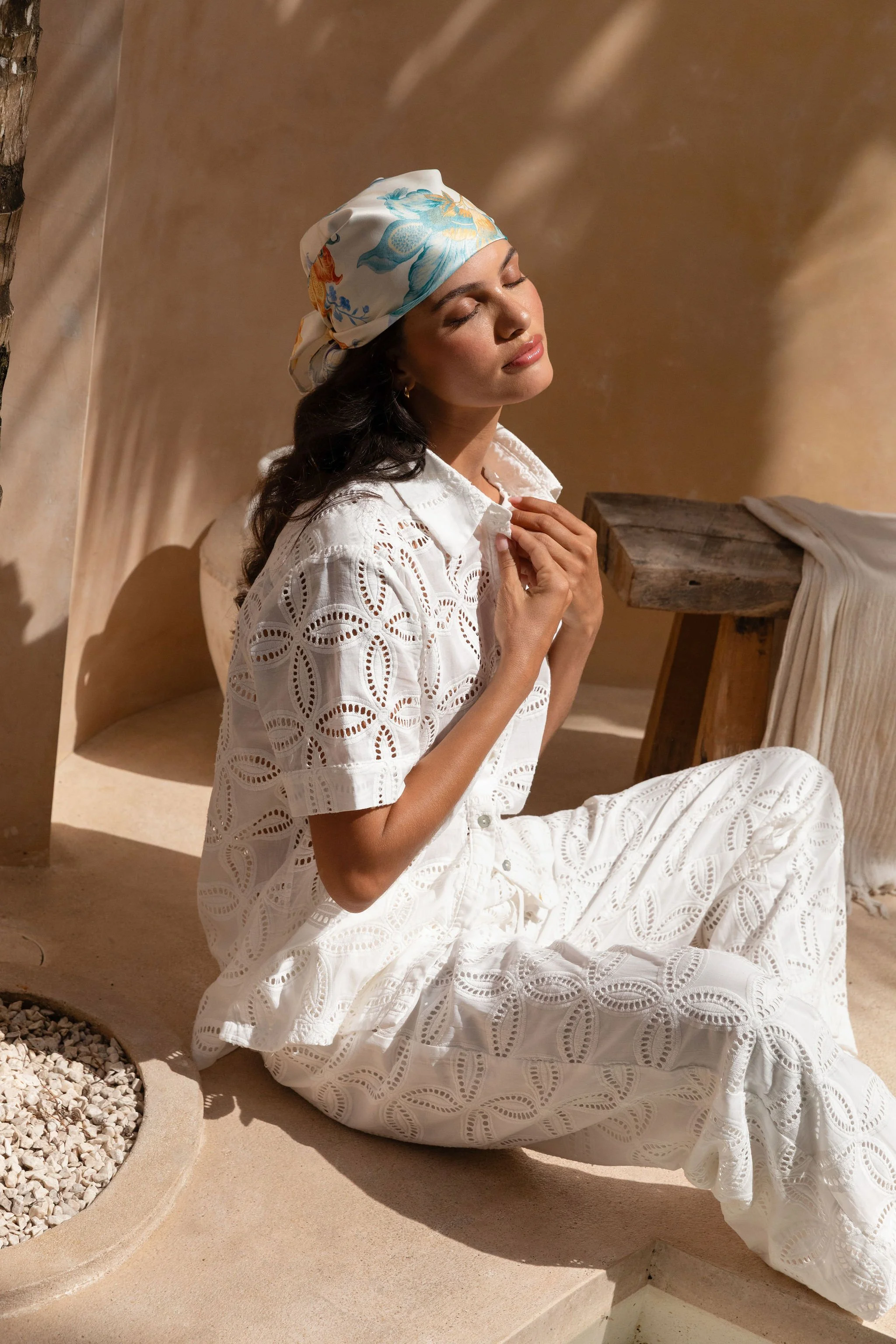 Woman in white eyelet dress and a colorful headscarf sitting with closed eyes, hands near her chest, in a sunlit outdoor setting.