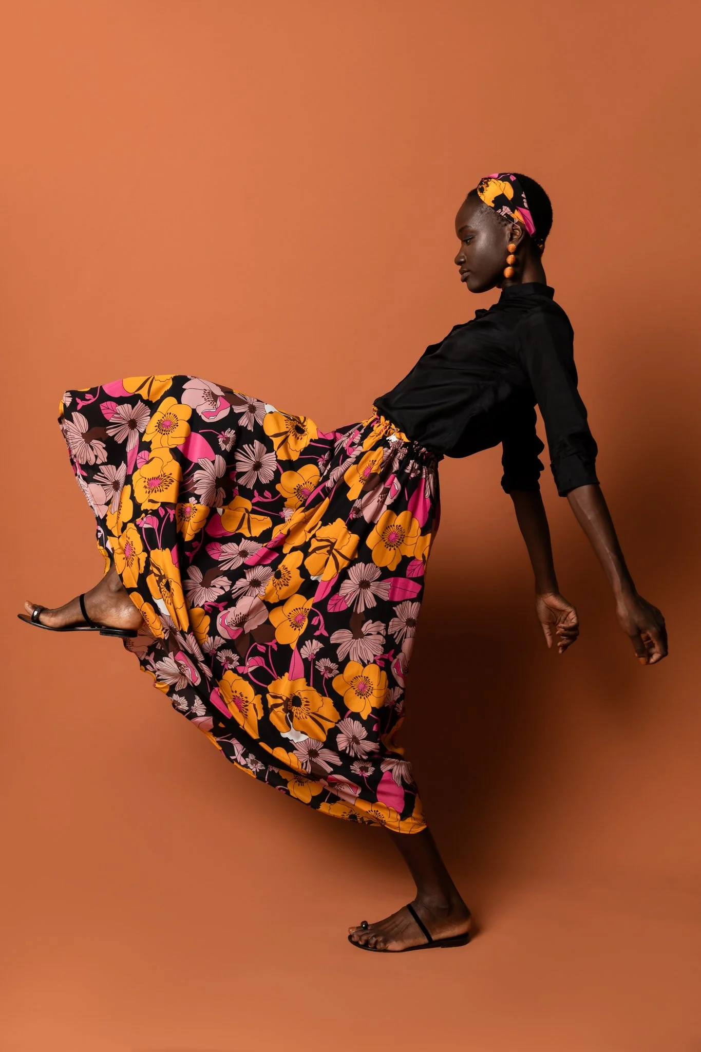 A woman with dark skin is posed reclining on a brown background. She is dressed in a black button-up top and a colorful floral skirt with shades of orange, pink, and white. She is wearing black sandals and has a pink, orange, and black headscarf and 