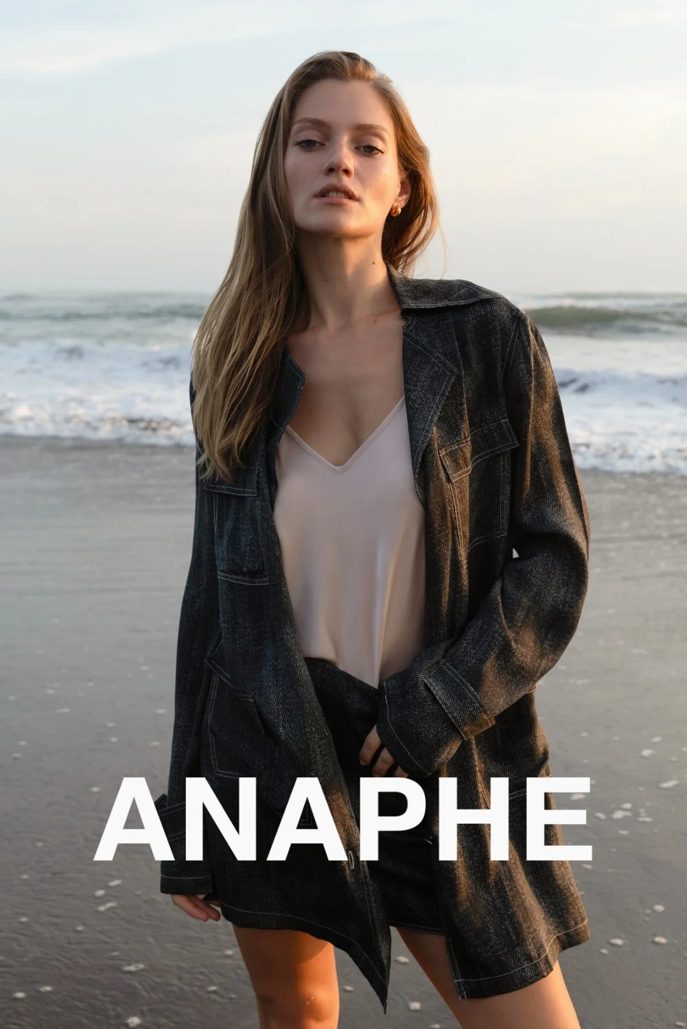 A young woman with long, light brown hair standing on a beach during sunset, wearing a black denim jacket over a beige top with the word 'ANAPHE' in bold white letters at the bottom.