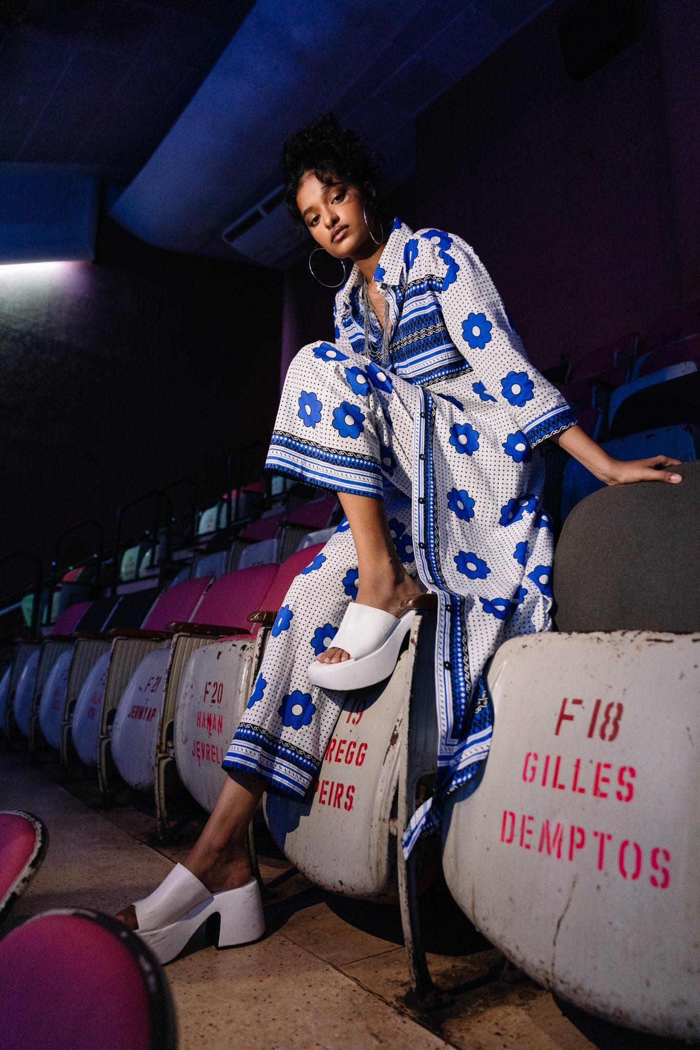 A young woman with dark curly hair, wearing a blue and white floral and striped pajama set, sitting on a row of stadium seats in a dimly lit arena. She is wearing large hoop earrings, silver necklaces, and white platform shoes. The seats behind her a