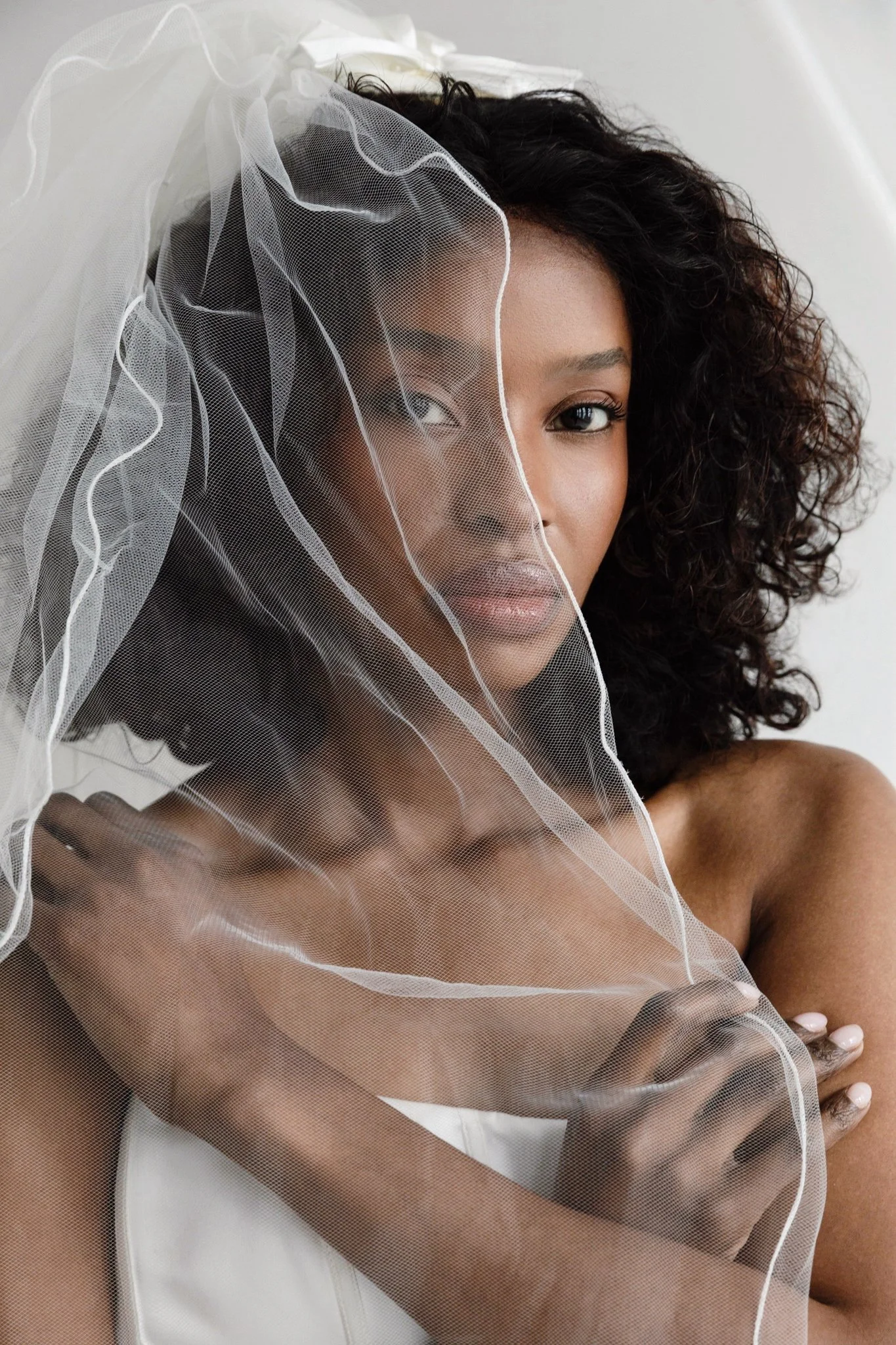 A woman with curly black hair wearing a sheer veil over her face and shoulder, with a white top, against a white background.