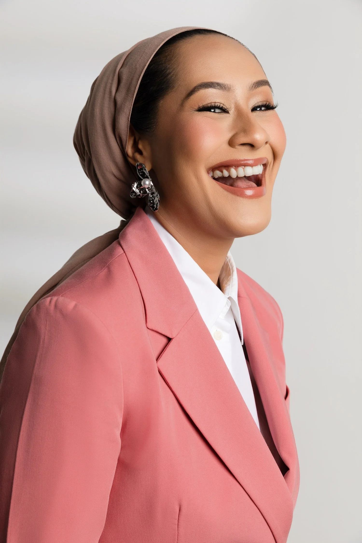 A smiling woman wearing a pink blazer, white shirt, beige headscarf, and large decorative earrings.