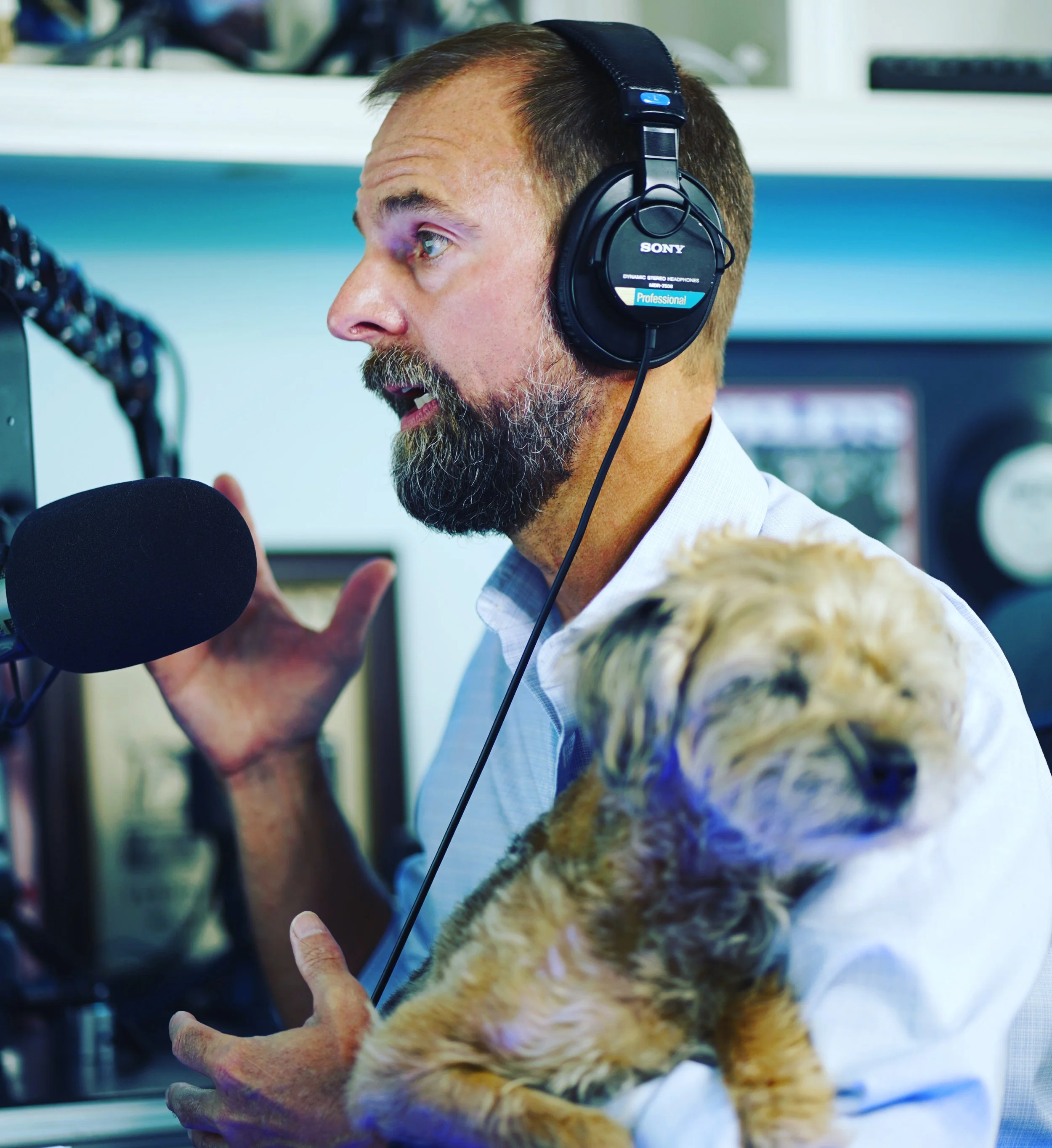 A man with a beard and short brown hair wearing Sony professional over-ear headphones talking into a microphone, with a dog in the foreground.