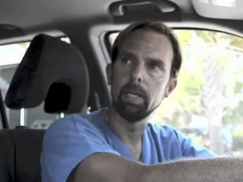 A man with a beard and mustache wearing a blue shirt sitting in a car, looking to his left.