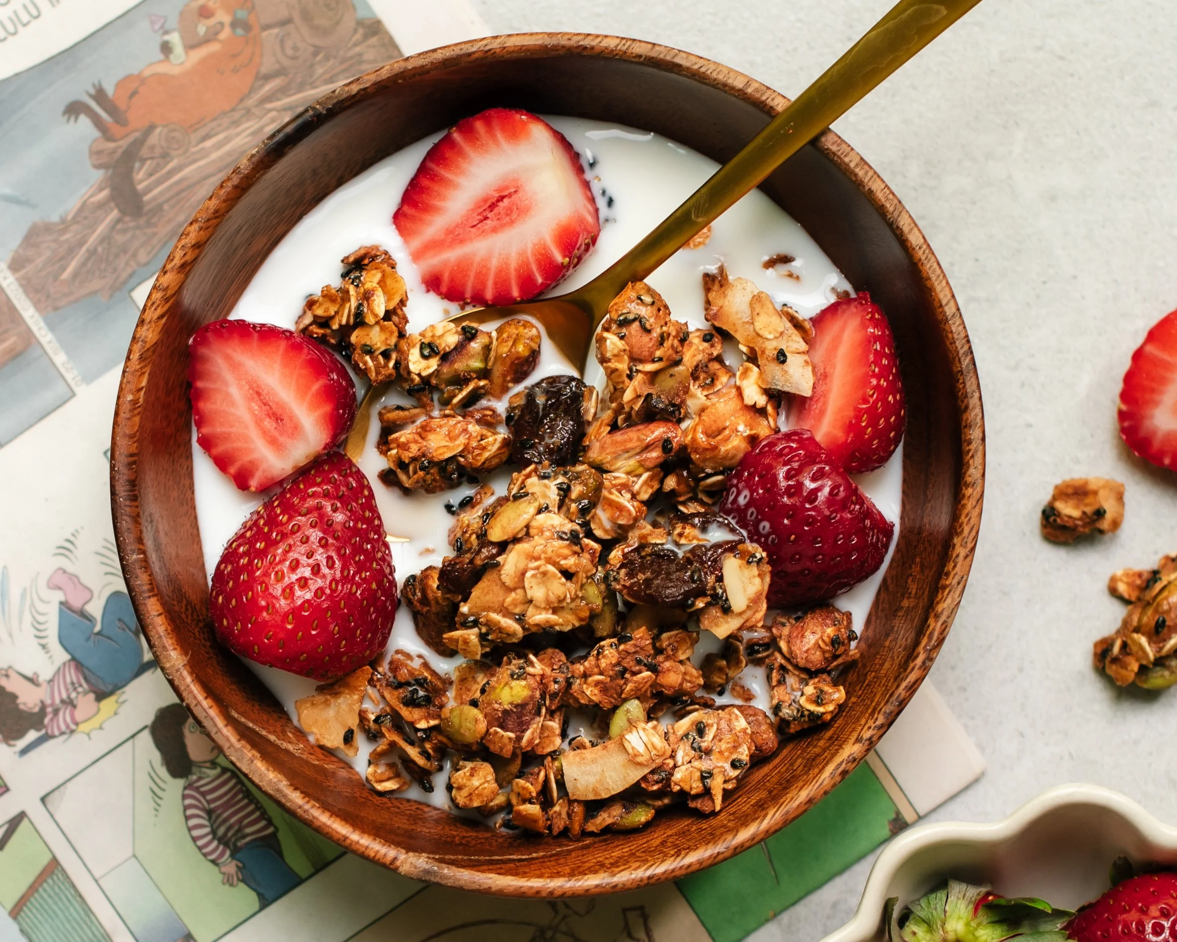 small wooden bowl of granola with fresh strawberries in milk on top of a light surface with comic strip partially in view