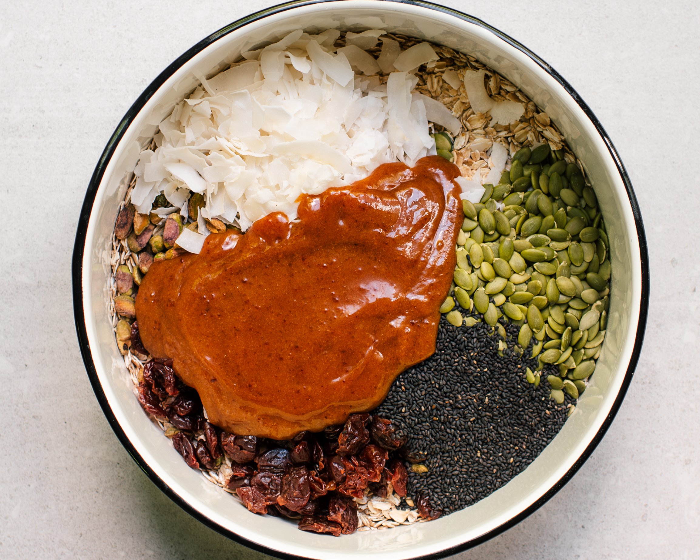 Large white mixing bowl containing granola ingredients oats, pumpkin seeds, dried cherries, pistachios, black sesame seeds and coconut chips with a thick brown sauce centered on top before being stirred.