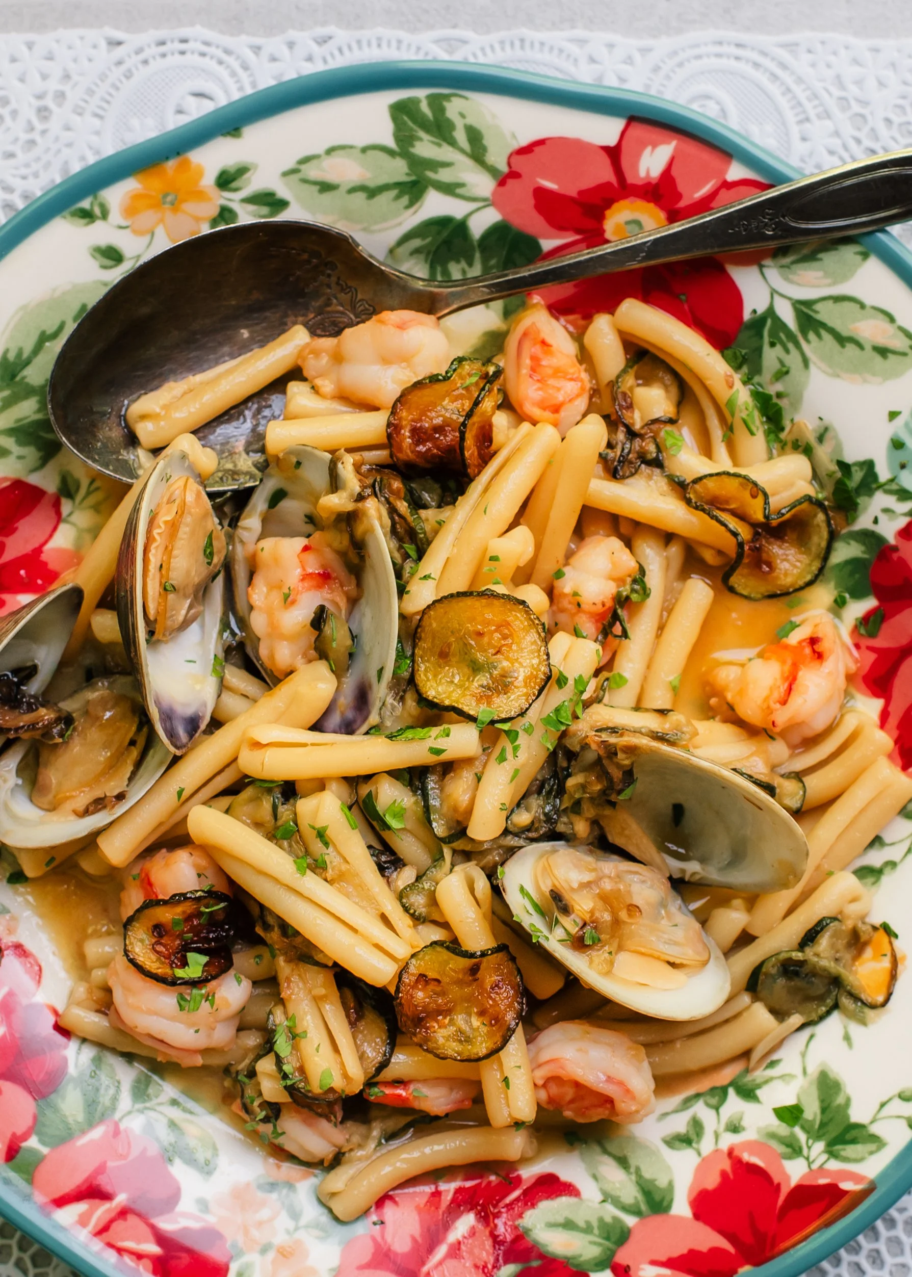 Fresh clams in shells, zucchini coins and pieces of shrimp tossed with short pasta on a colorful serving plate set on top of a lace surface.