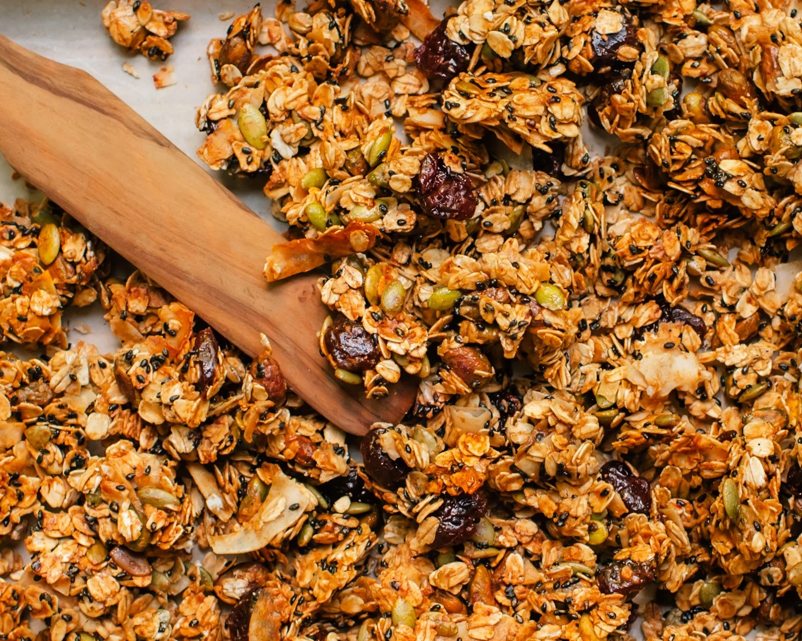 close up of granola with dried cherries, coconut chips, pistachios and pumpkin seeds on a sheet pan with a wooden spatula.