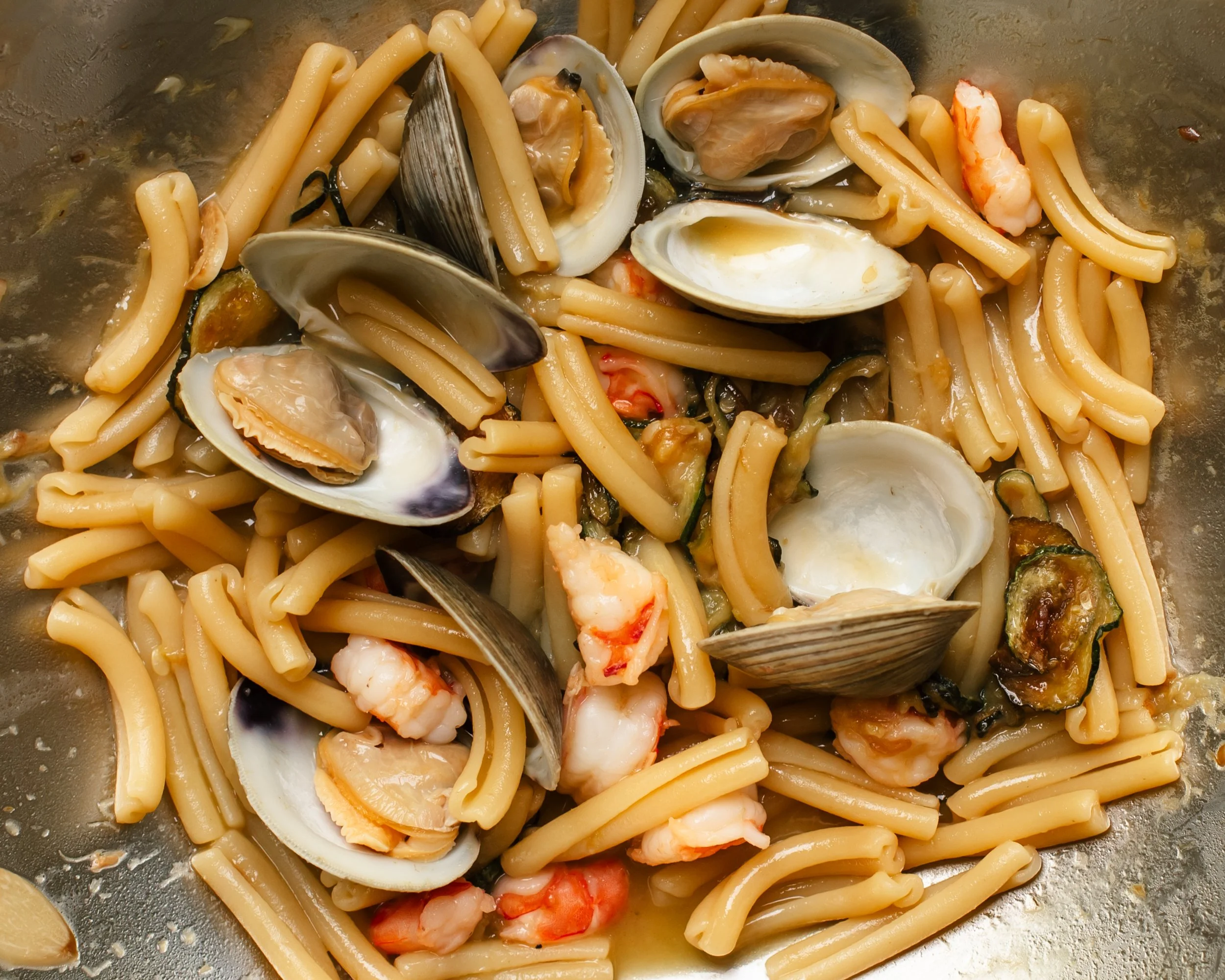 Fresh clams in shells, zucchini coins and pieces of shrimp tossed with short pasta in a metal pan.
