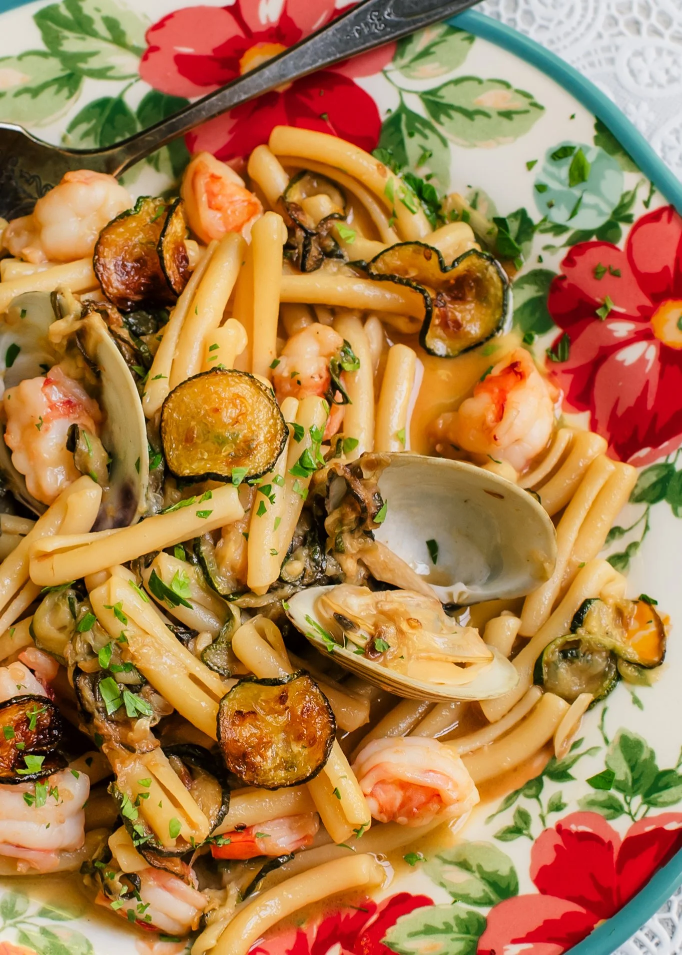 Fresh clams in shells, zucchini coins and pieces of shrimp tossed with short pasta on a colorful serving plate set on top of a lace surface.
