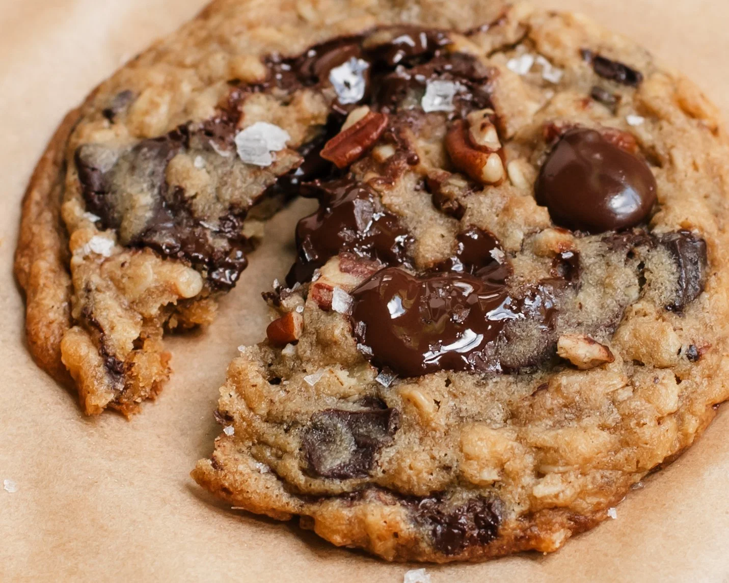 Single cookie dotted with chocolate chips, small pecan pieces and flakey salt on beige parchment paper