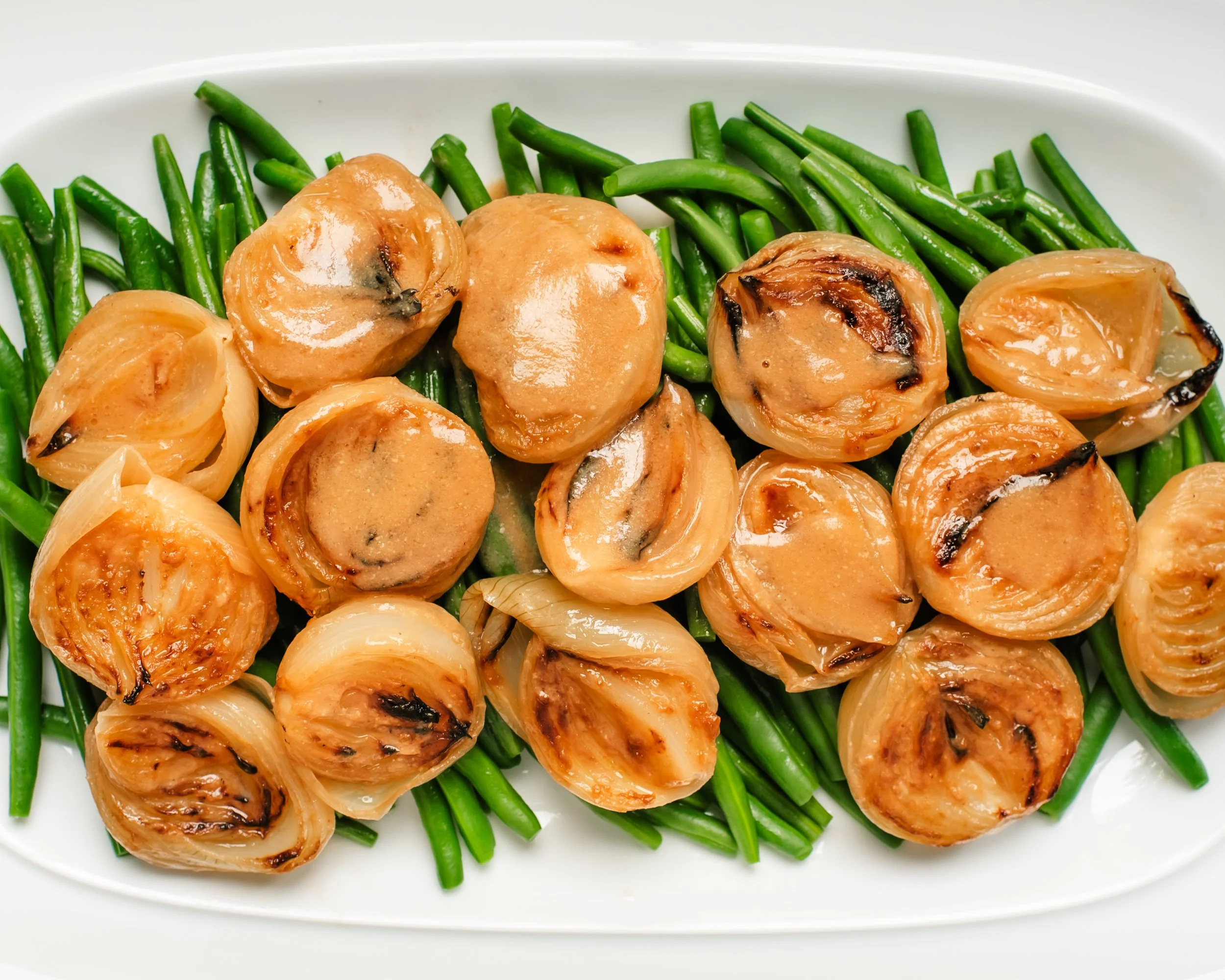 Roasted white onion halves covered in sauce on top of blanched green beans on white platter
