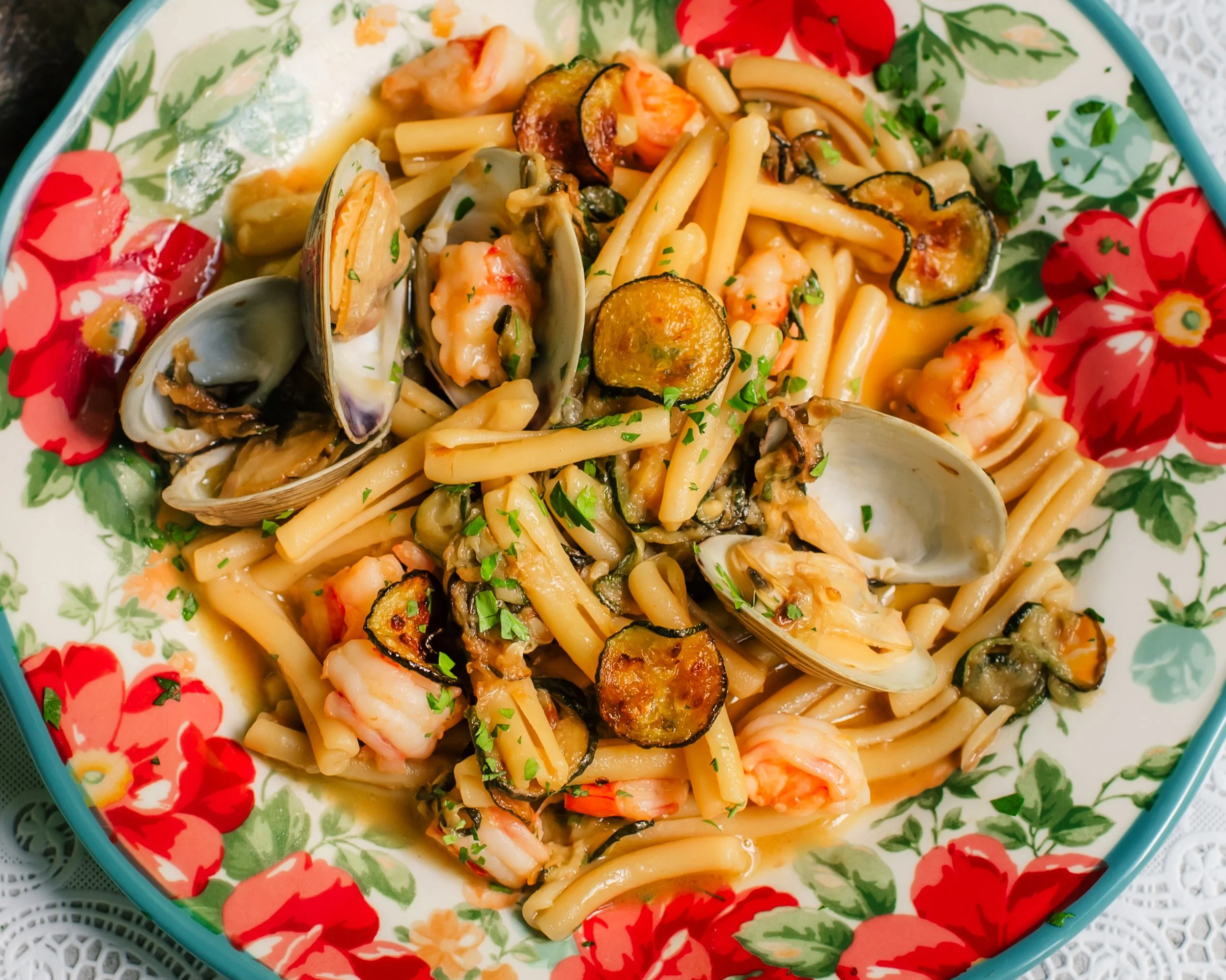 Fresh clams in shells, zucchini coins and pieces of shrimp tossed with short pasta on a colorful serving plate set on top of a lace surface.