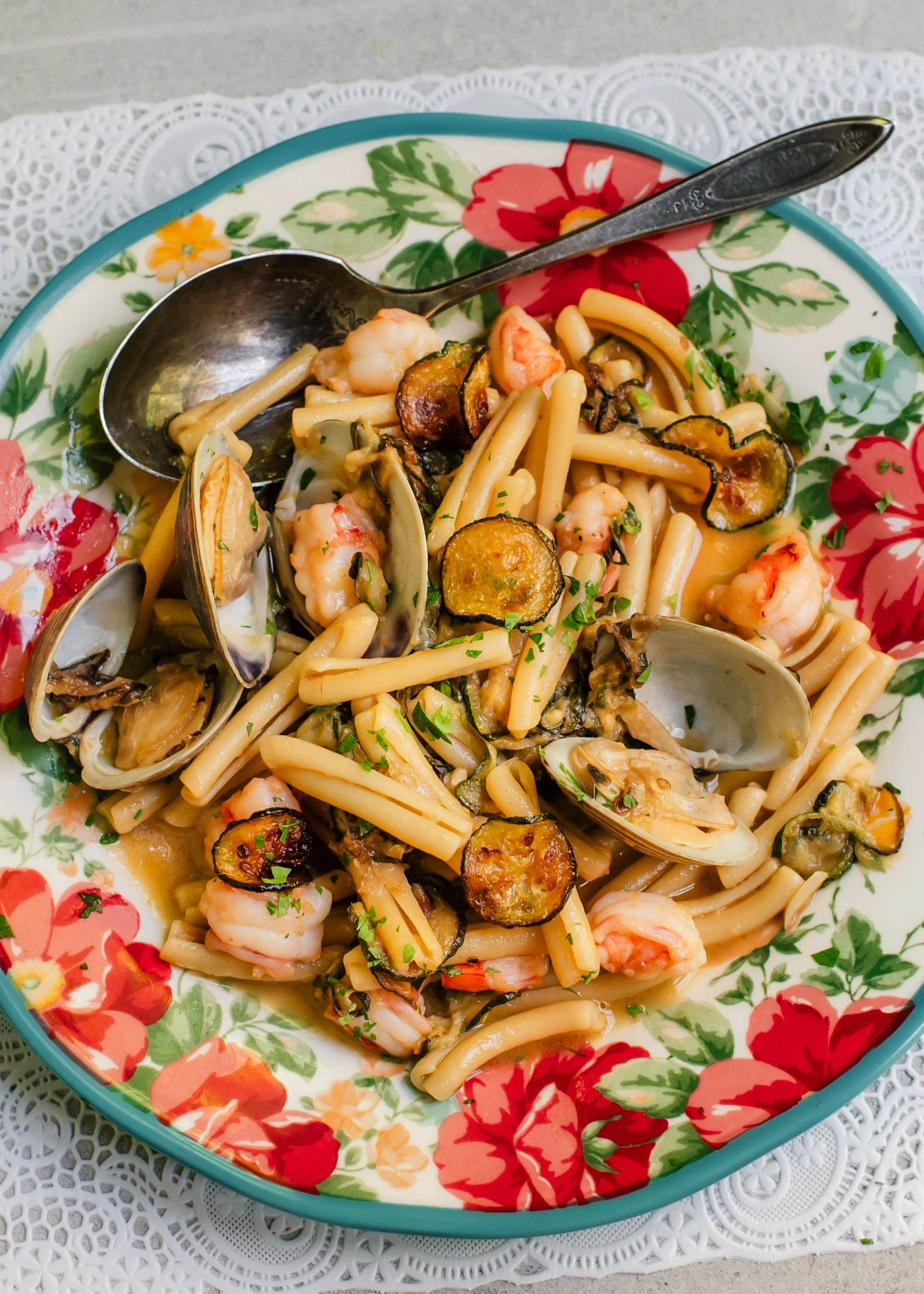 Fresh clams in shells, zucchini coins and pieces of shrimp tossed with short pasta on a colorful serving plate set on top of a lace surface.