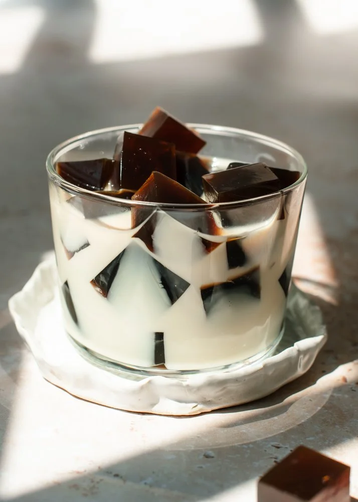 Glass bowl containing coffee jelly with milk, placed on a white plate, on a surface with natural light.