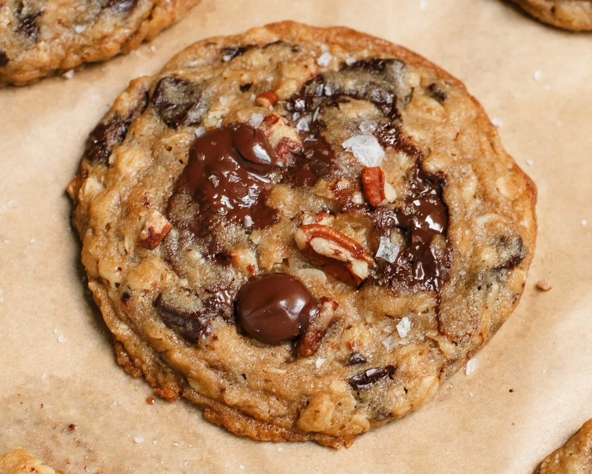 Single cookie dotted with chocolate chips, small pecan pieces and flakey salt on beige parchment paper