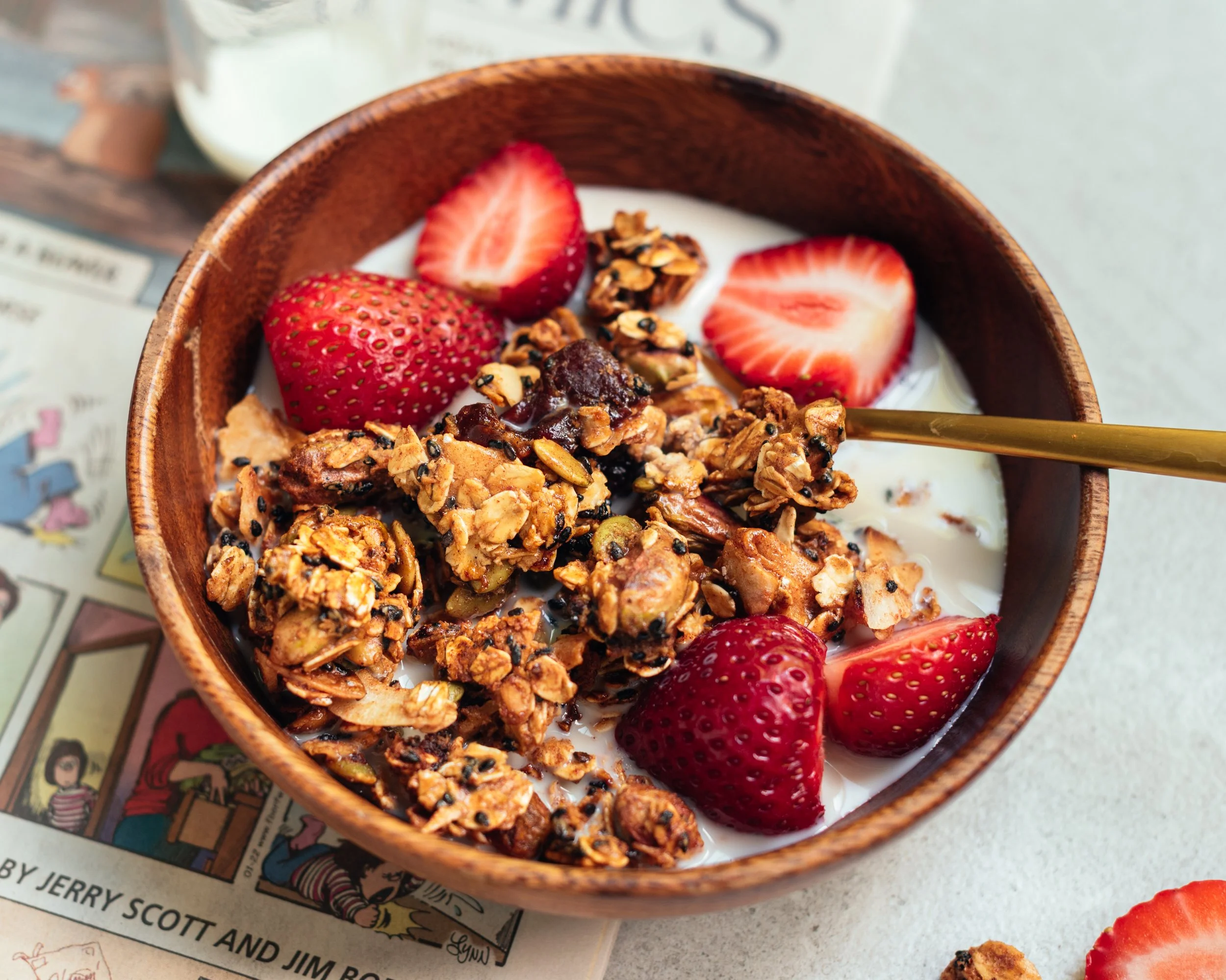 small wooden bowl of granola with fresh strawberries strawberries in milk wth a golden spoon, on light coloured surface with partial view of comic strip