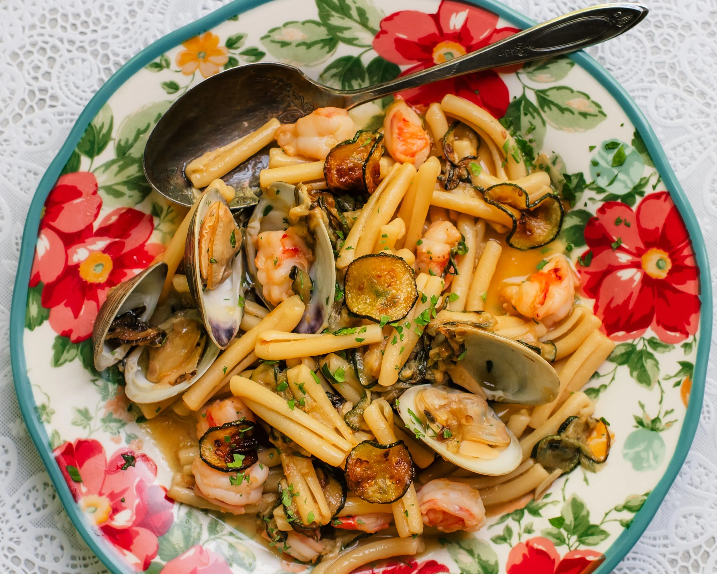 Fresh clams in shells, zucchini coins and pieces of shrimp tossed with short pasta on a colorful serving plate set on top of a lace surface.
