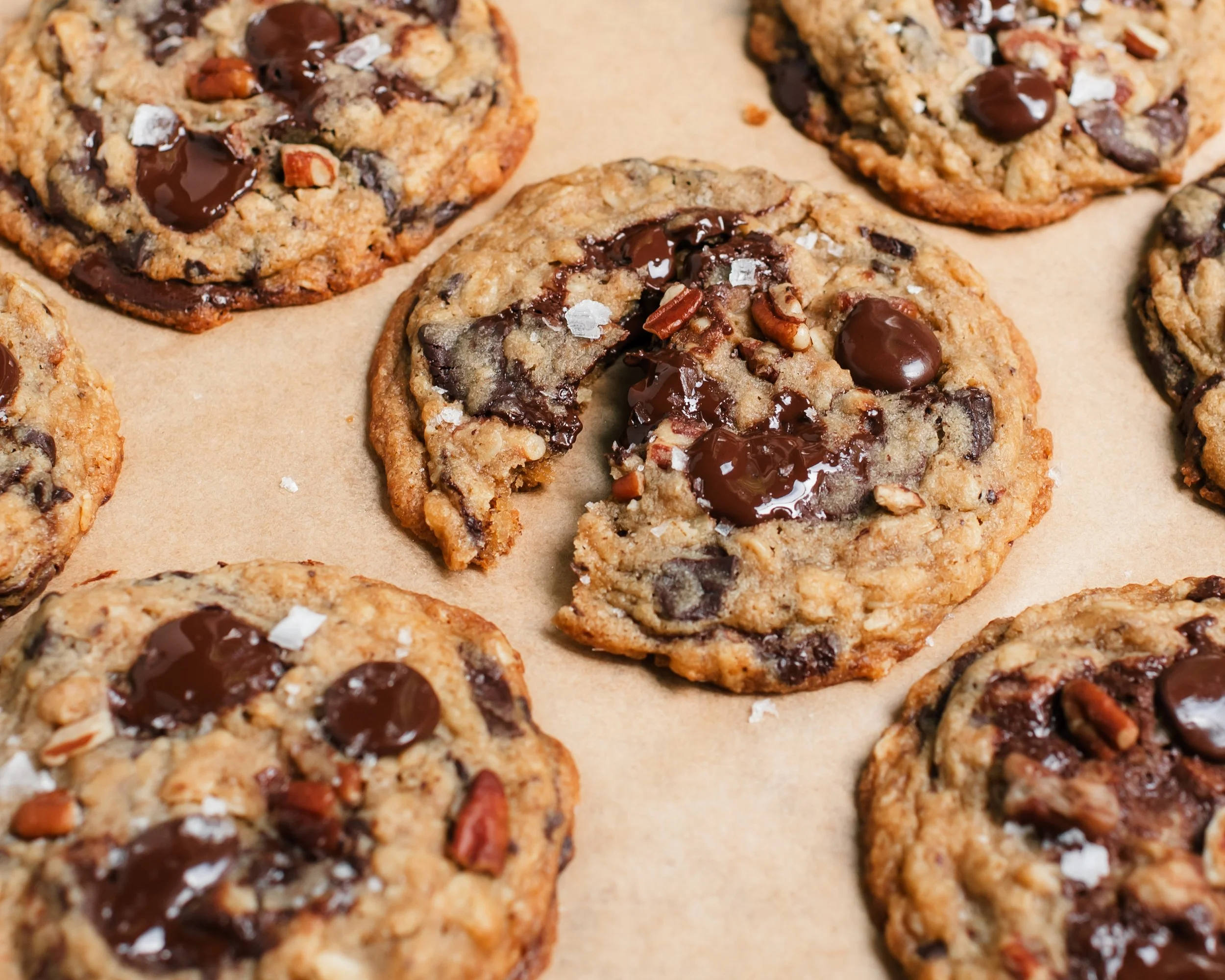 Cookies dotted with chocolate chips, small pecan pieces and flakey salt on beige parchment paper