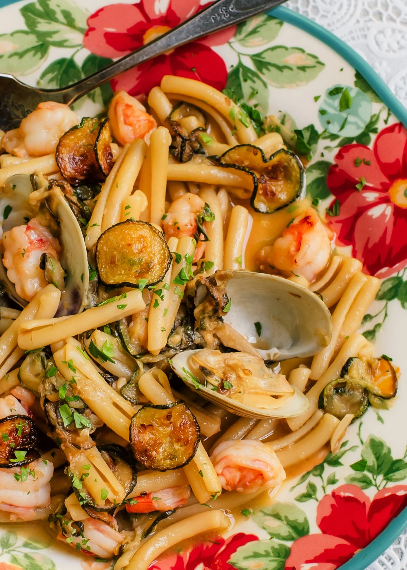 Fresh clams in shells, zucchini coins and pieces of shrimp tossed with short pasta on a colorful serving plate set on top of a lace surface.