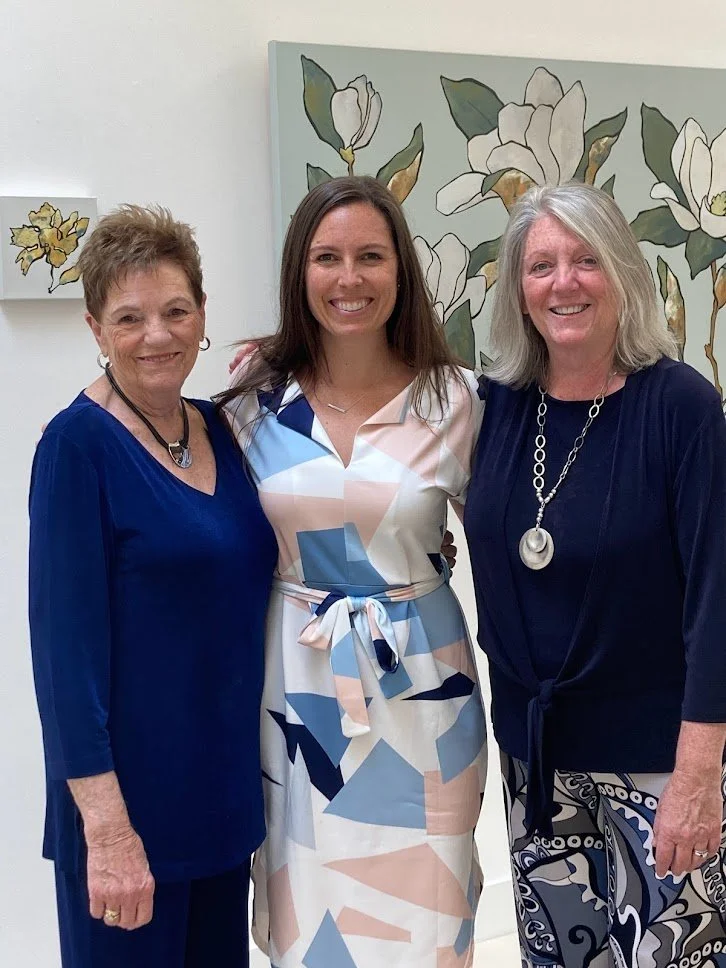 Three women standing together indoors, smiling at the camera. They are dressed in colorful and elegant outfits, with a floral painting in the background.