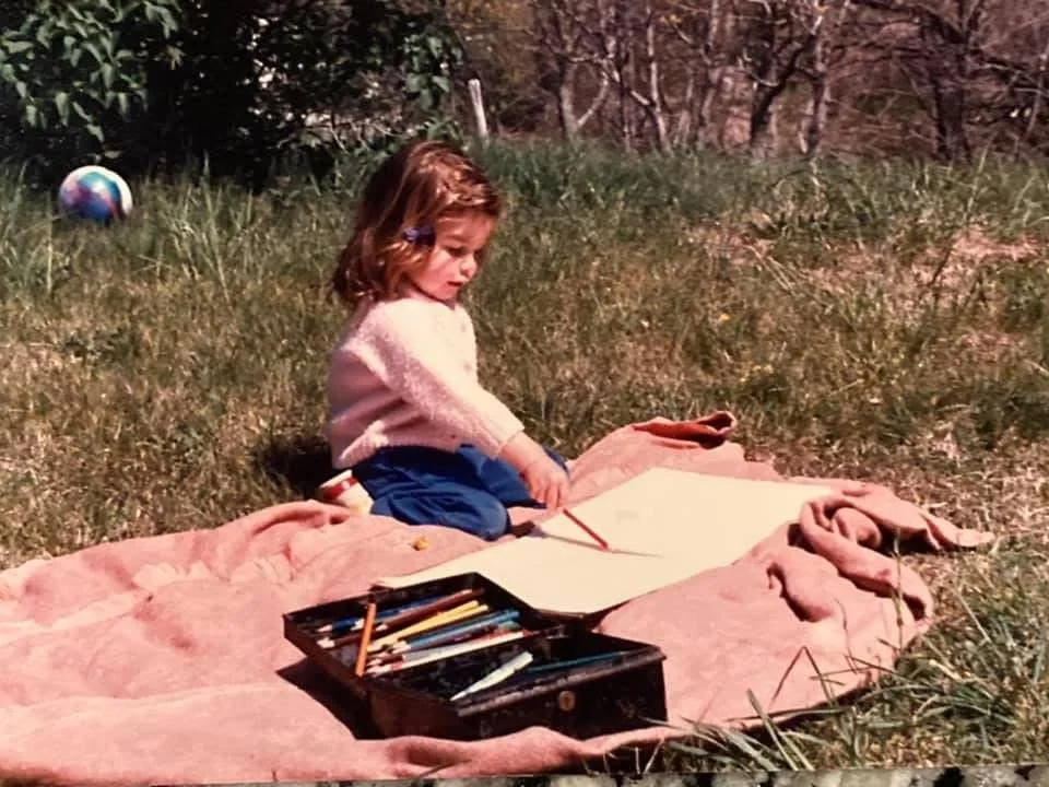 Young Meg Davies with brown hair sitting on a pink blanket outdoors, drawing with colored pencils on a large sheet of paper. An open box of colored pencils is in front of her on the blanket, and a colorful ball is in the grass behind her.