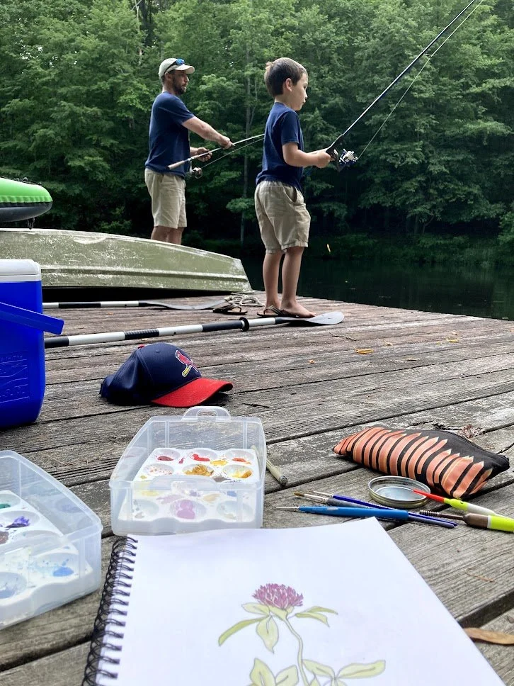 Two boys fishing off a wooden dock by a lake, with a boat and trees in the background. The dock has art supplies, a notebook, and a baseball cap on it.
