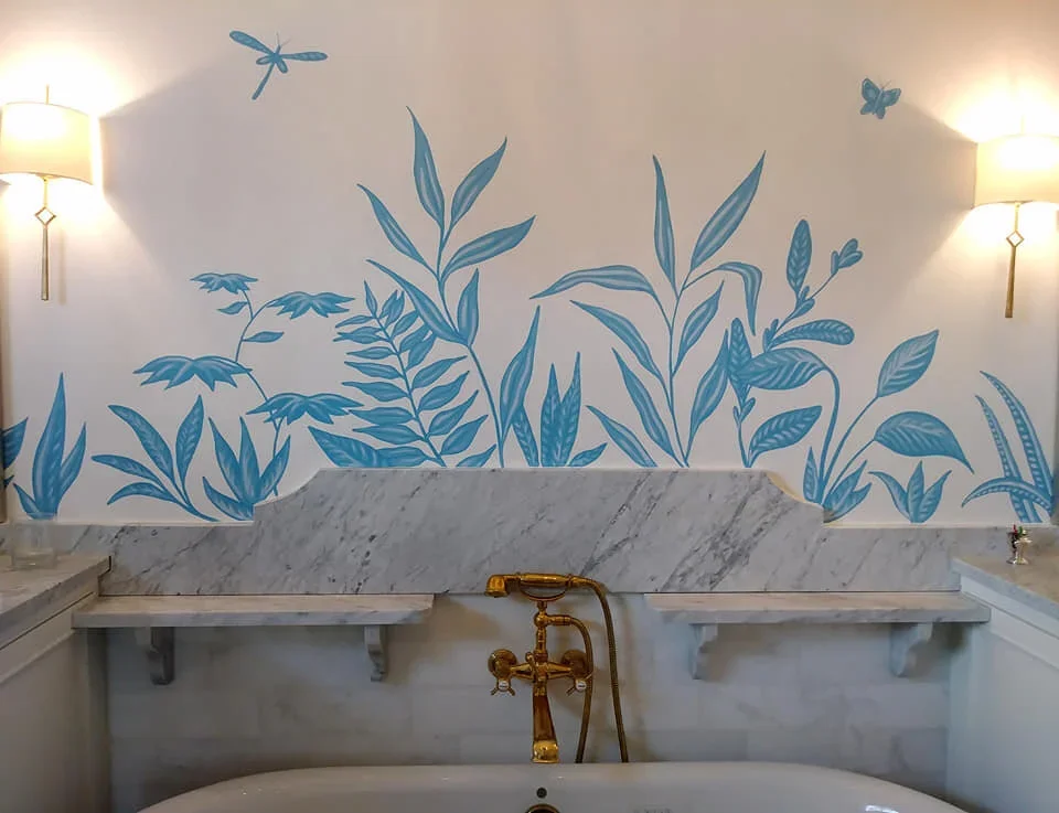 Bathroom with a white marble bathtub, gold faucet, and a wall mural of blue botanical and insect illustrations, illuminated by wall sconces.