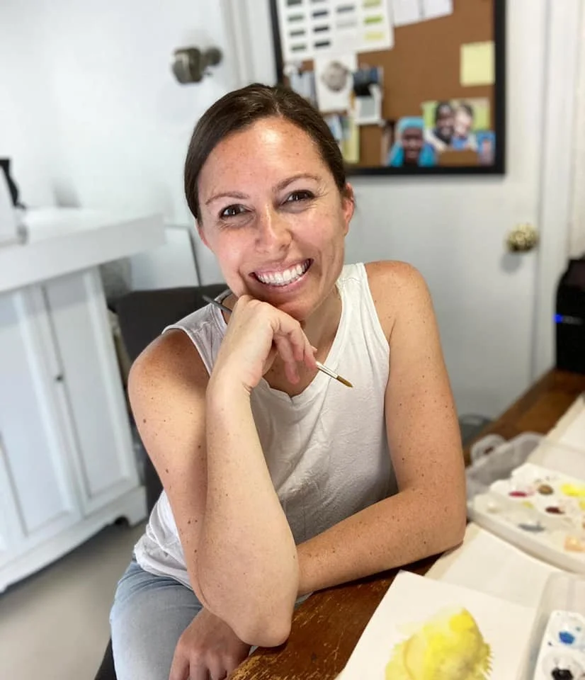 Artist Meg Davies with short brown hair and freckles, smiling and holding a paintbrush, sitting at a table with watercolor paints and paper.