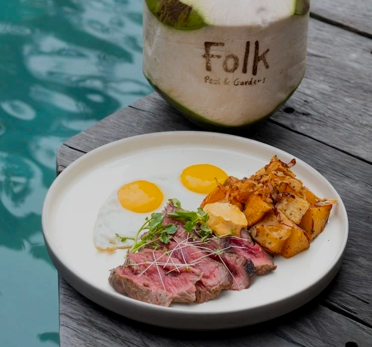 A breakfast plate with two fried eggs, a piece of cooked steak topped with microgreens, roasted potatoes, and a dollop of sauce, on a wooden table near a pool, and a coconut drink