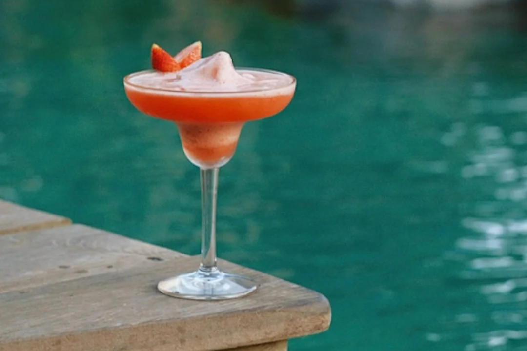 A pink cocktail with foam and a strawberry garnish in a stemmed glass, sitting on a wooden surface near a pool.