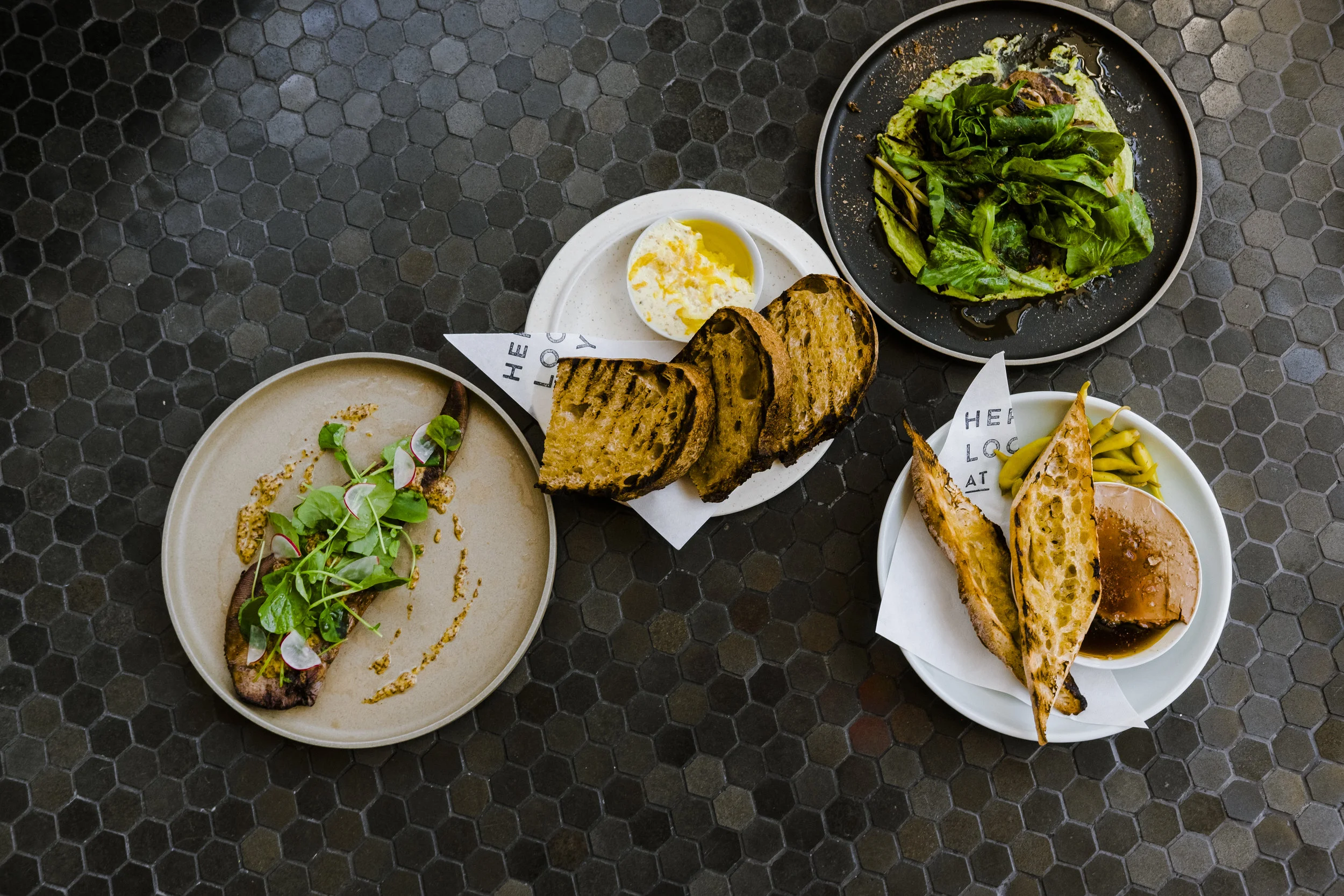 Image depicts Smoked beef tongue with garum mustard, cress, horseradish. Bub &amp; grandma’s bread with shiitake butter and sea salt. Lamb shoulder chop with miso, greens, minari, onion, and togarashi. And Chicken liver with basque peppers, smoked maple, and Bub &amp; Grandma's baguette.