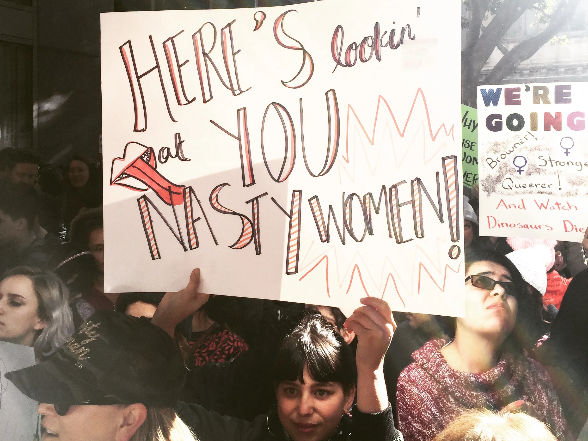 Image depicts a photo of a young woman holding a sign depicting the words “Here’s lookin’ at you NASTY WOMEN!” with a drawing of red lips and a tongue.