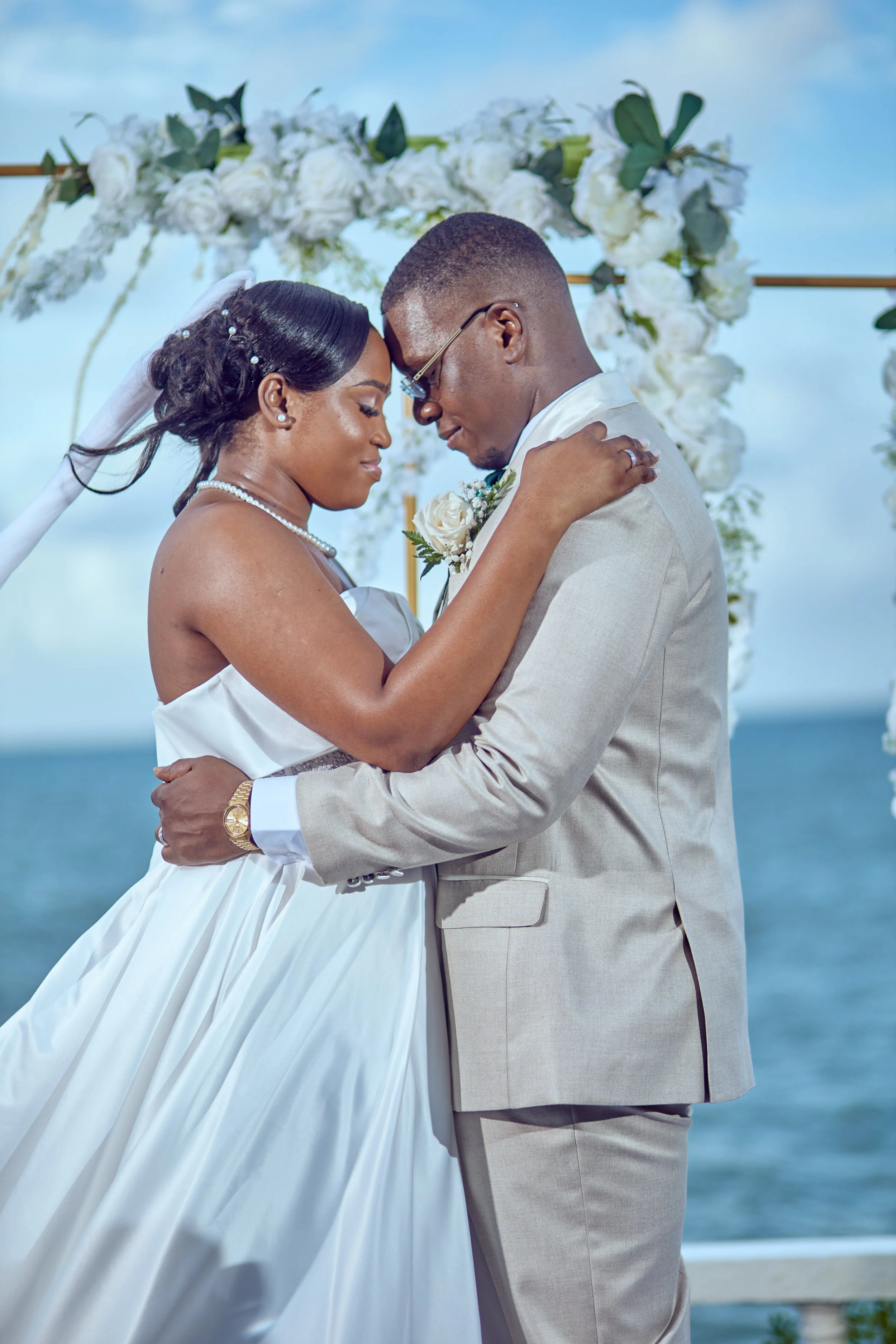 A bride and groom embracing at their outdoor wedding ceremony near the ocean, with a floral arch in the background.