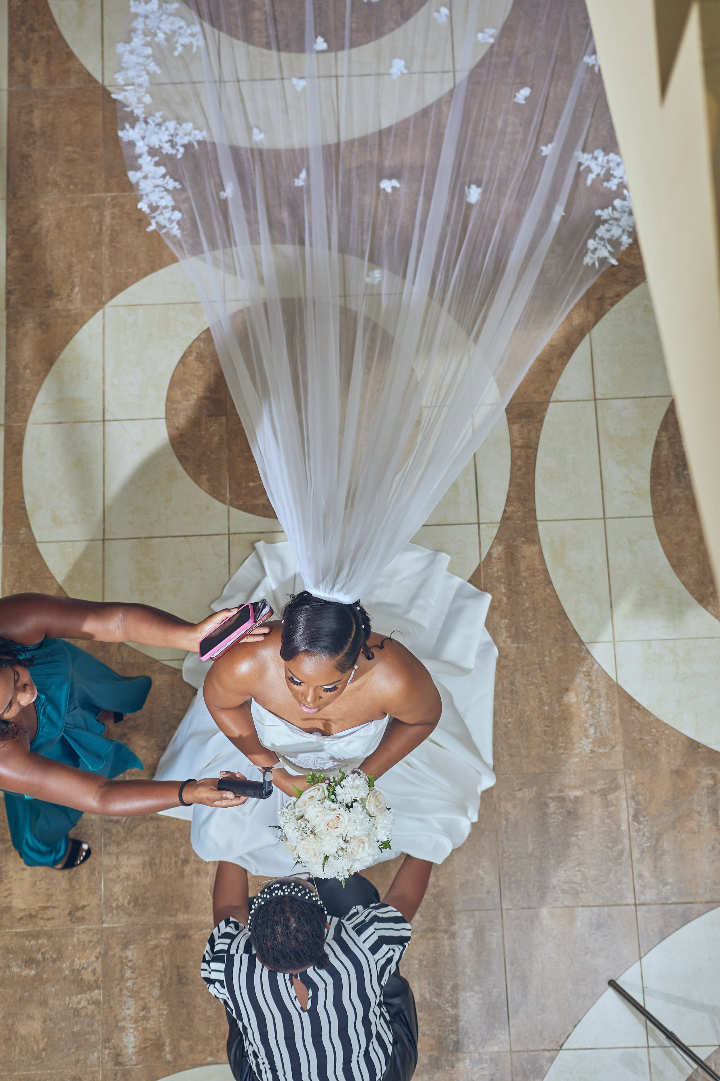 Overhead view of a bride in a white strapless wedding gown holding a bouquet of white flowers, standing on a patterned tile floor, as her wedding officiant, wearing a black and white striped top, stands in front of her. Two women are beside her, one 