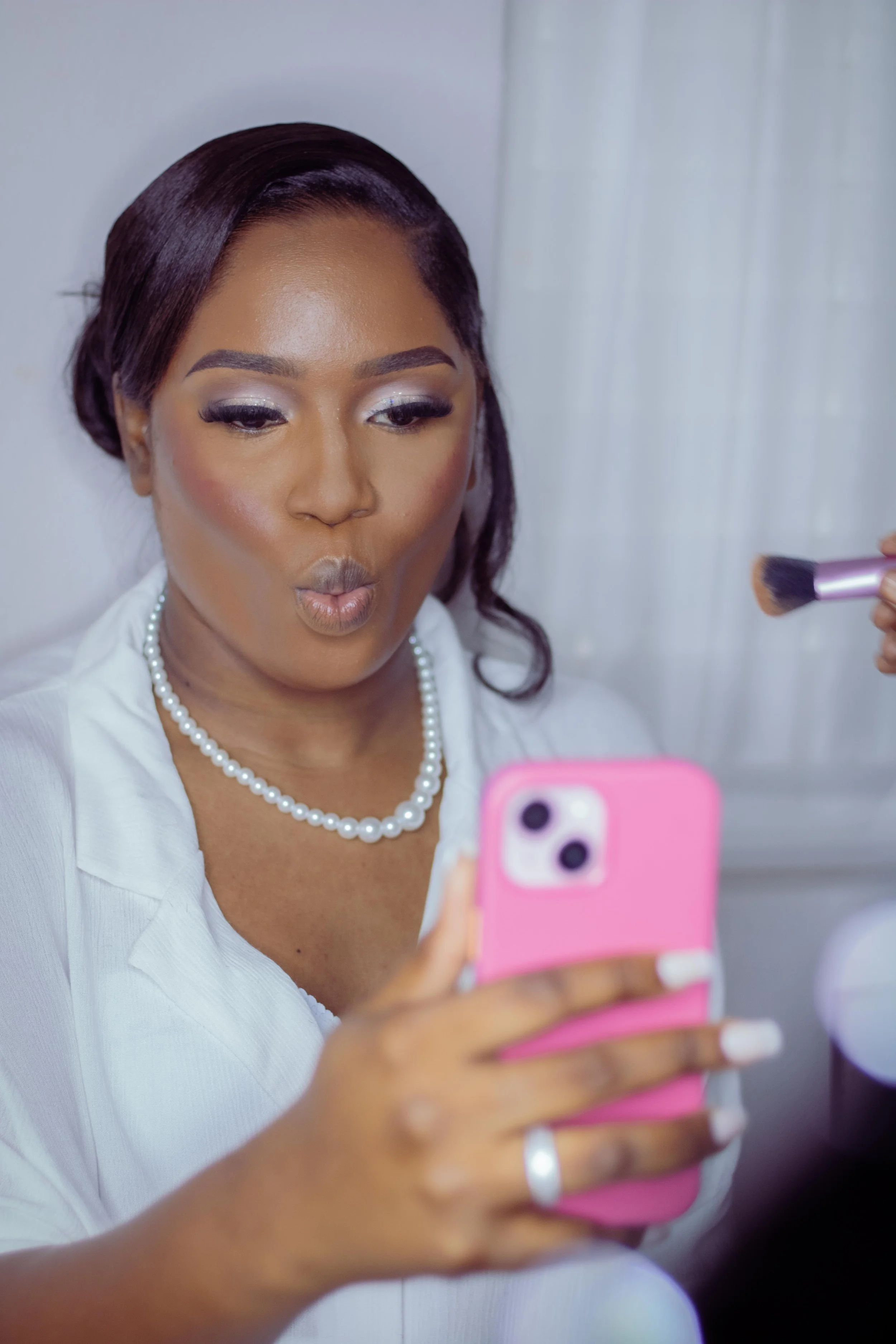 A woman with makeup and a pearl necklace is taking a selfie with her pink phone, while someone is applying makeup with a brush.