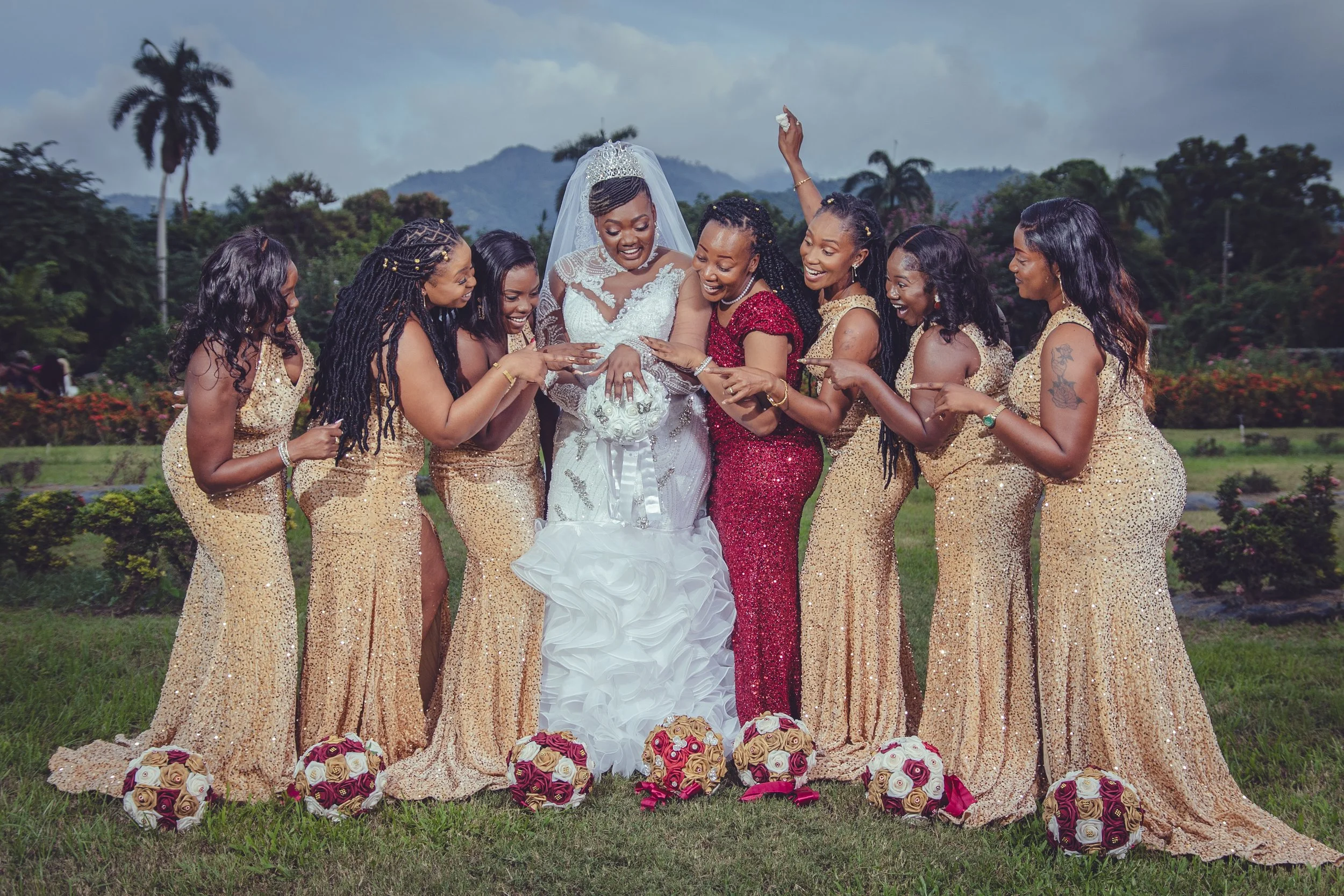 A bride in a white wedding gown and veil celebrates with her bridesmaids outside in a garden, surrounded by bouquets of roses, with mountains and palm trees in the background.