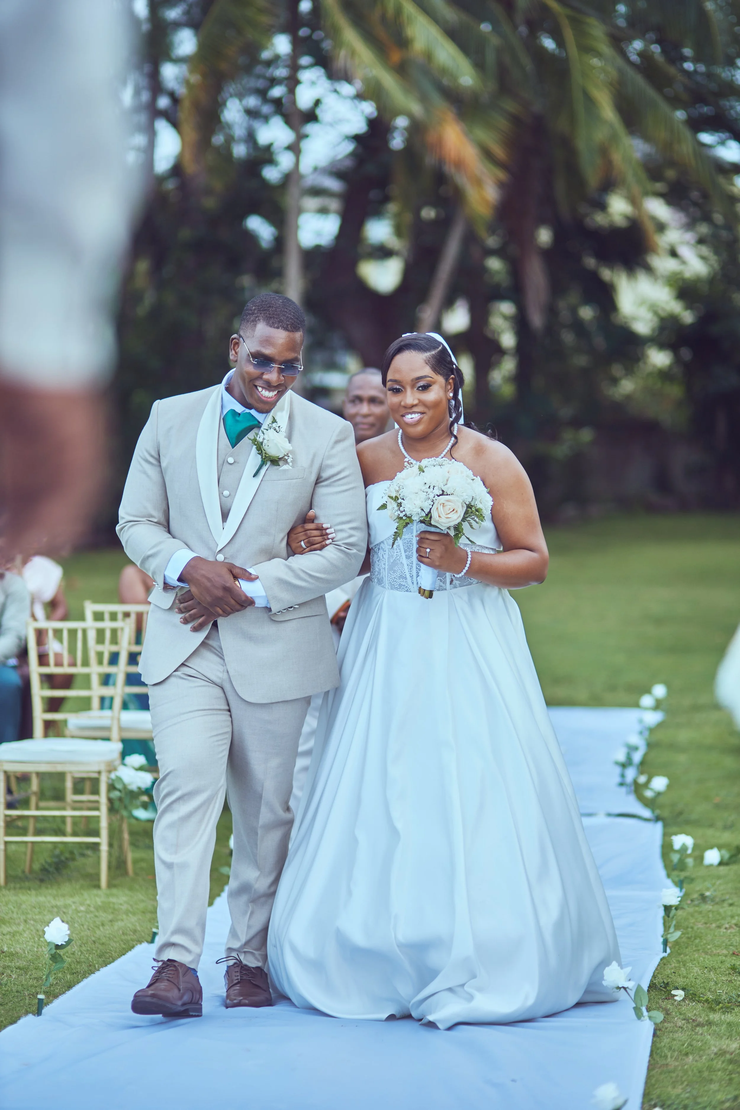 A bride and groom walking down the aisle during a wedding ceremony outdoors surrounded by trees, with the bride holding a bouquet of white flowers and the groom wearing a light-colored suit and sunglasses.