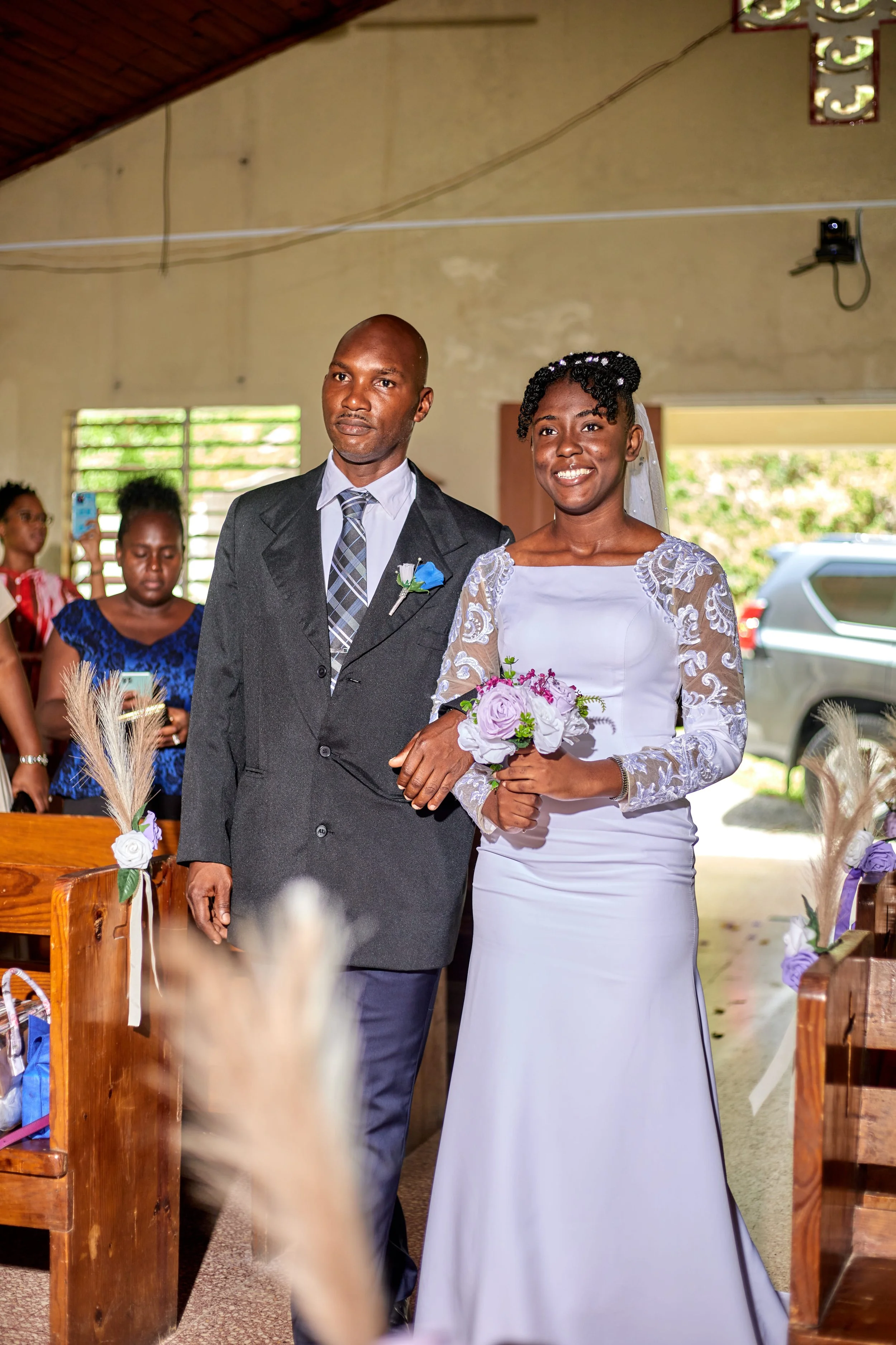 A bride in a white wedding dress holding a bouquet walks arm in arm with a groom in a dark suit inside a church or wedding venue.
