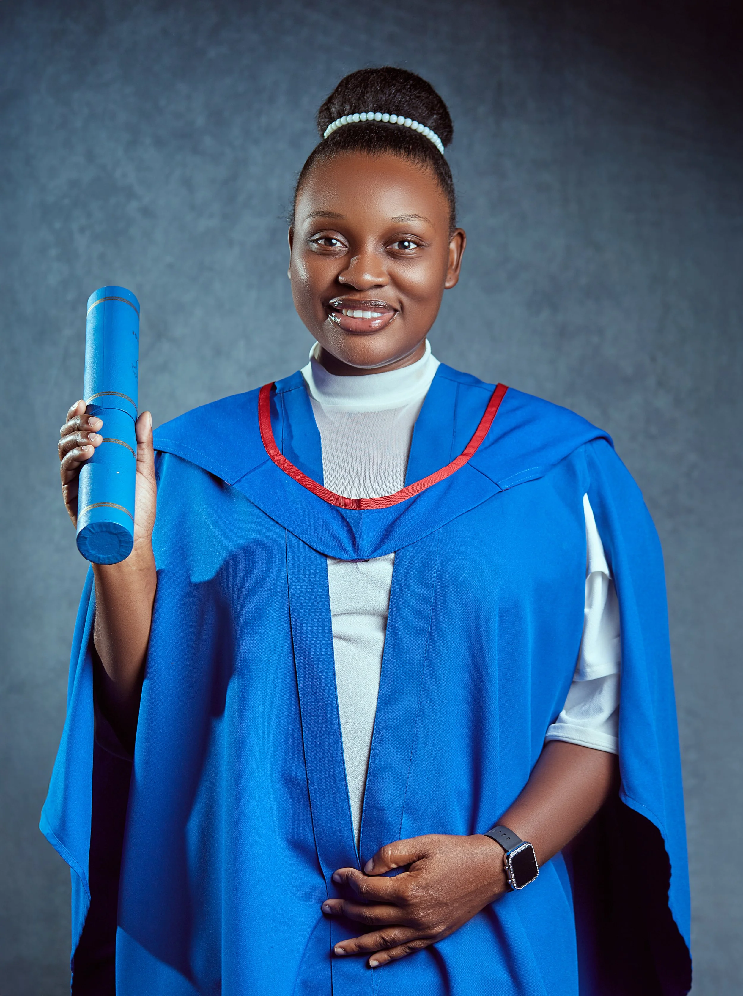 Young woman in graduation gown holding a rolled diploma, standing against a dark gray background.