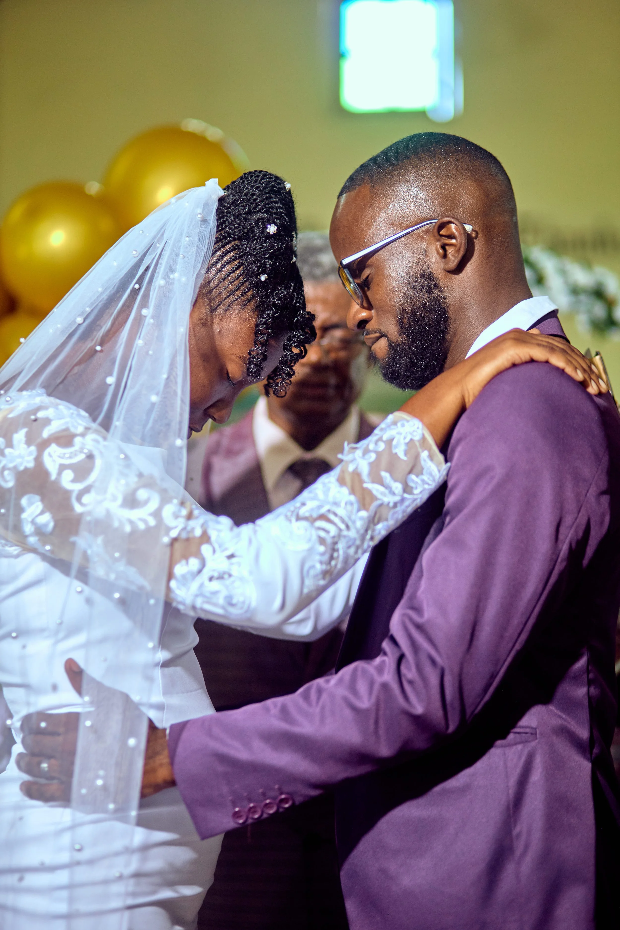 A couple kneels with foreheads touching during a wedding ceremony, with a officiant and golden balloons in the background.