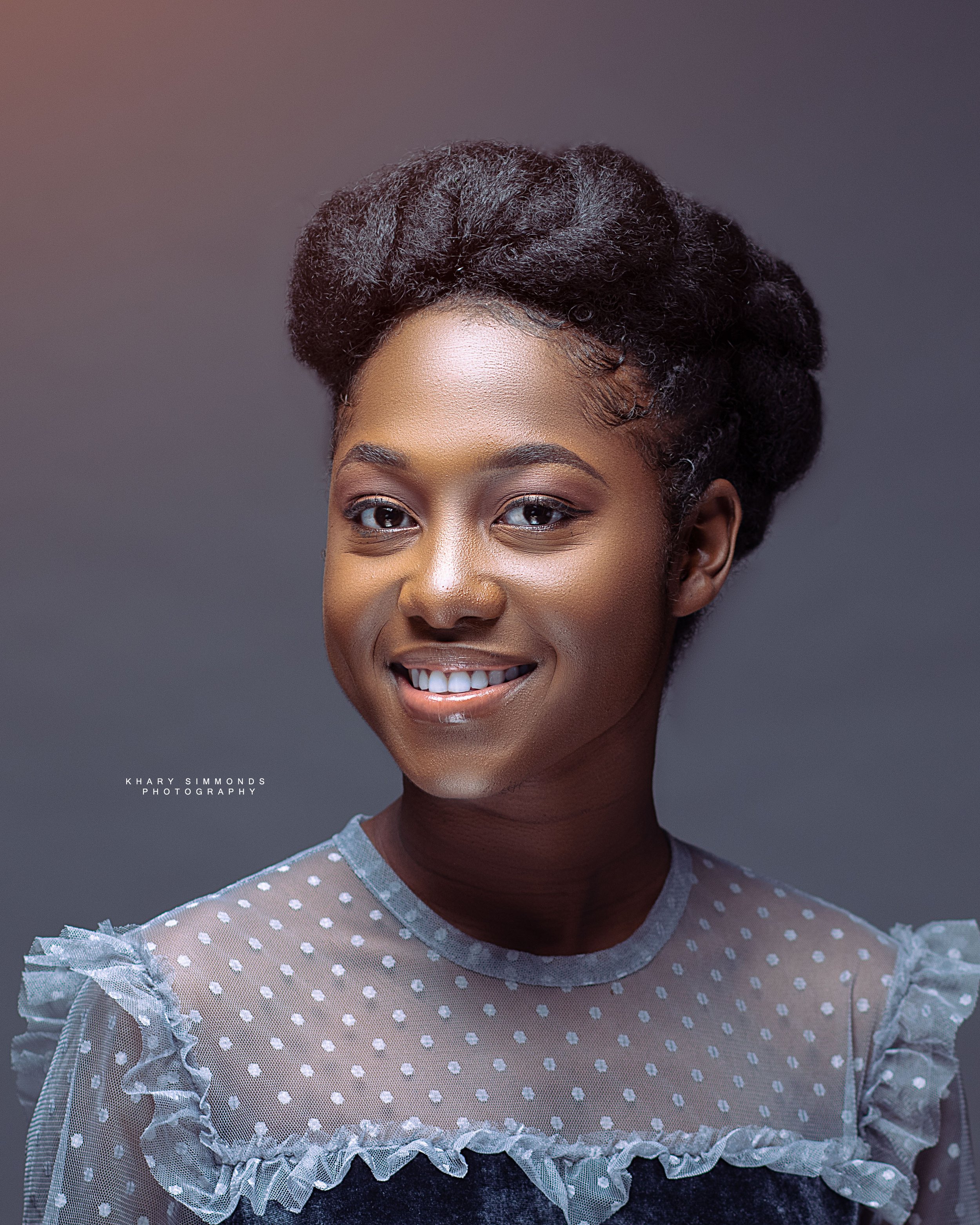 A young woman with dark hair styled in an updo and a smile, wearing a sheer polka dot blouse with ruffled details.