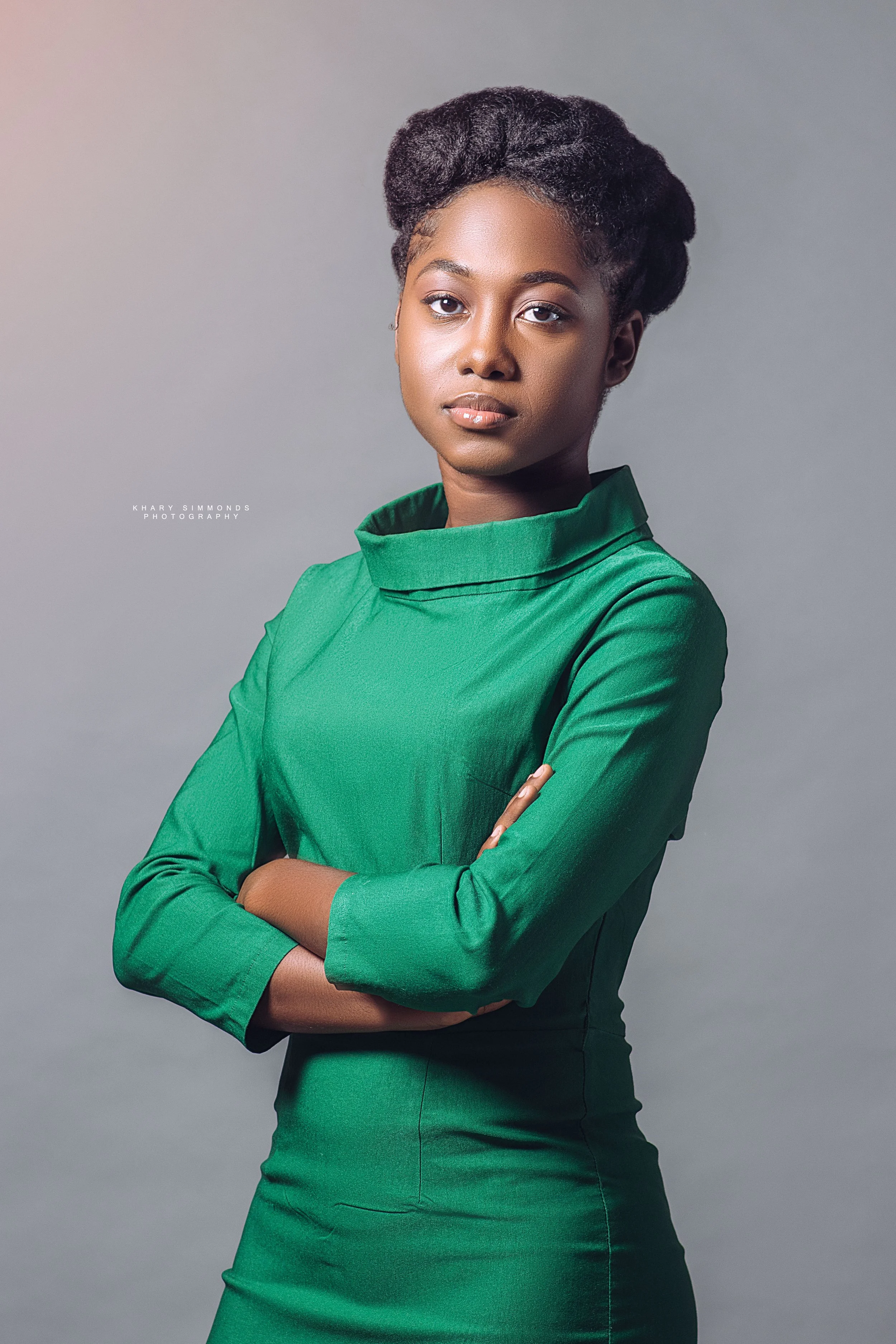 A young woman with short, dark, styled hair, wearing a green dress with a high collar, standing with her arms crossed against a plain gray background.