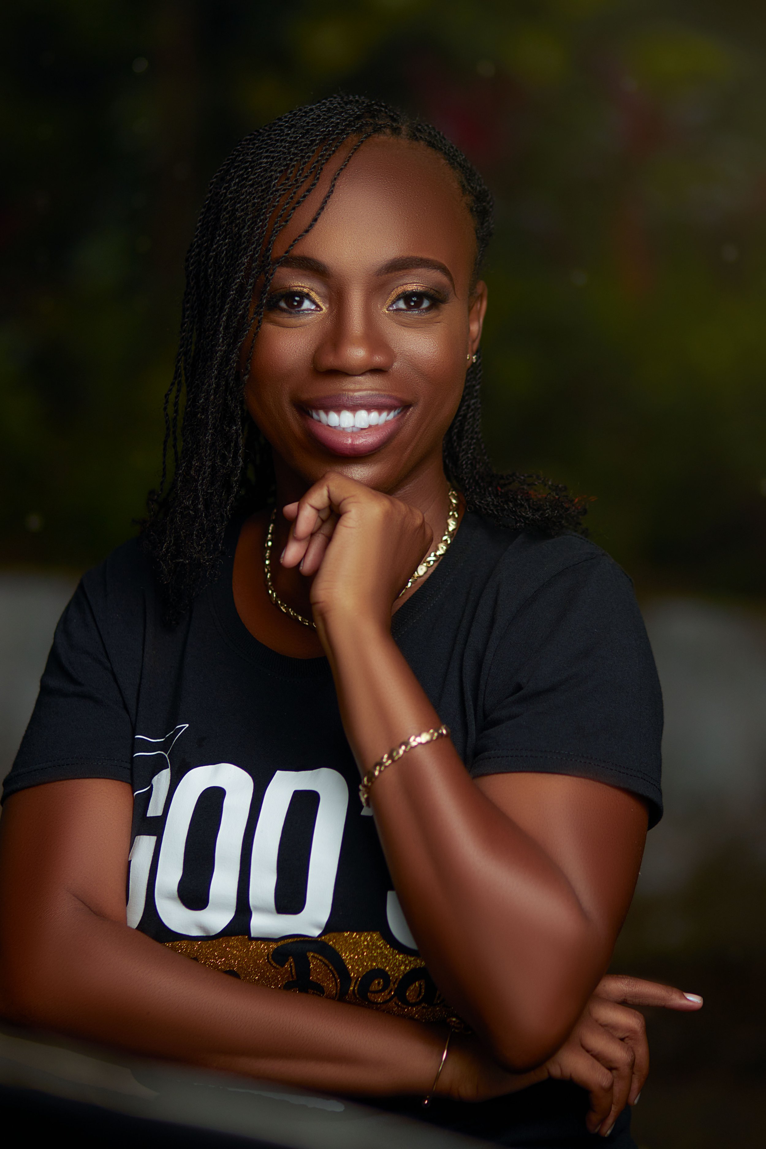 A woman with braided hair smiling, wearing a black T-shirt, a gold necklace, and bracelets, outdoors during dusk.