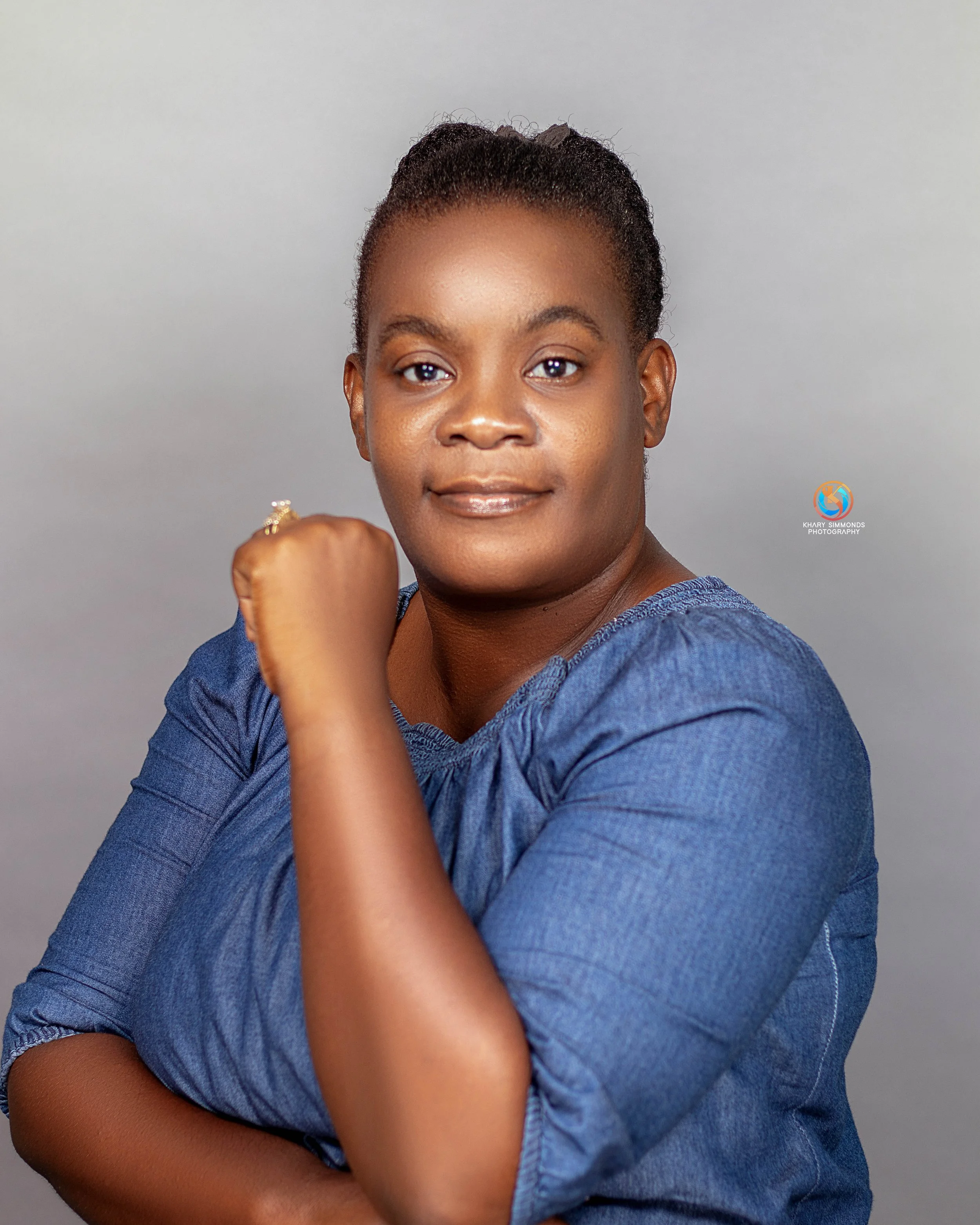 Portrait of a woman wearing a blue dress, posing with her arm bent and fist near her chin, against a plain gray background.