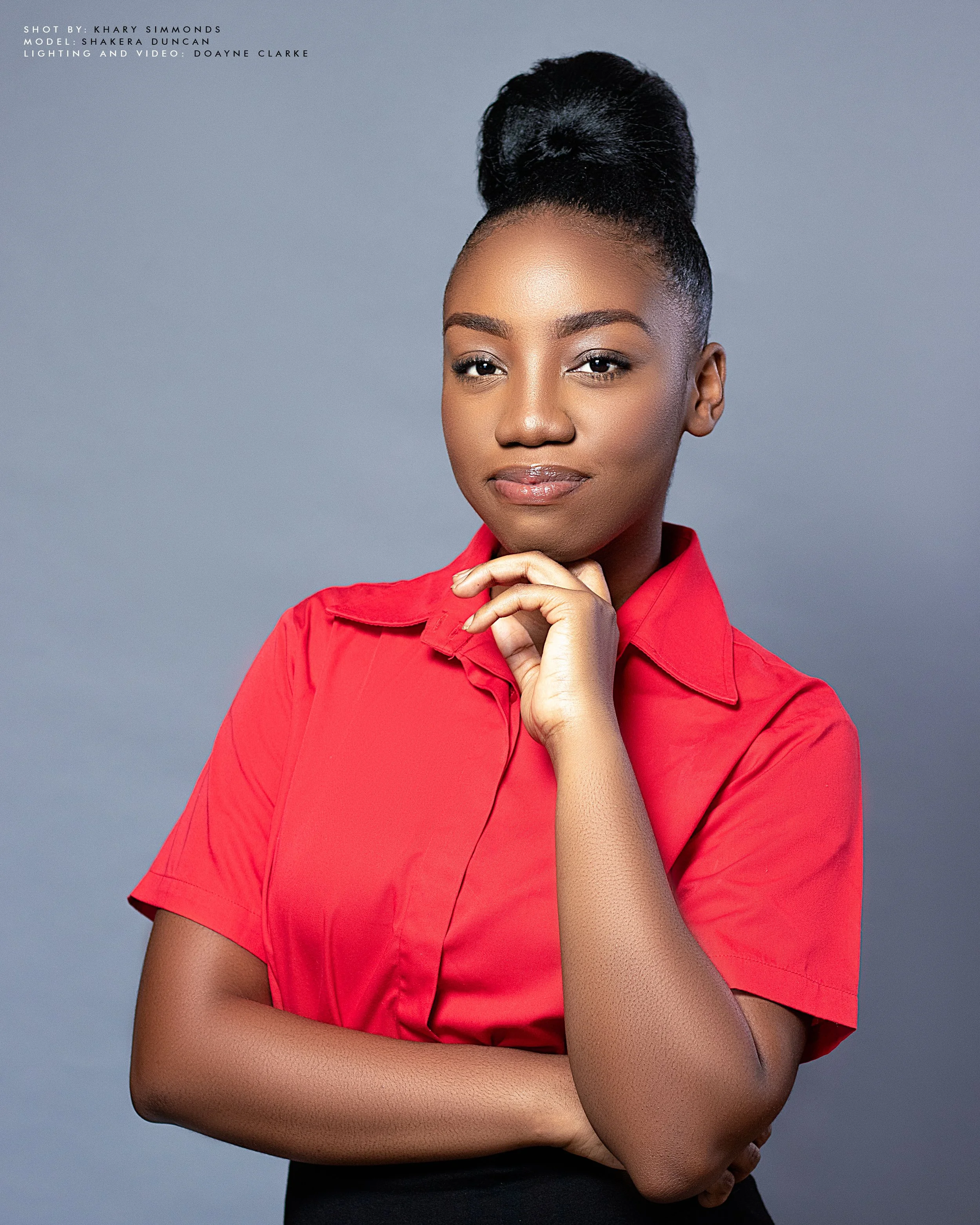 Portrait of a woman with dark hair in an elegant updo, wearing a bright red collared shirt, posing confidently with her chin resting on her hand against a plain gray background.