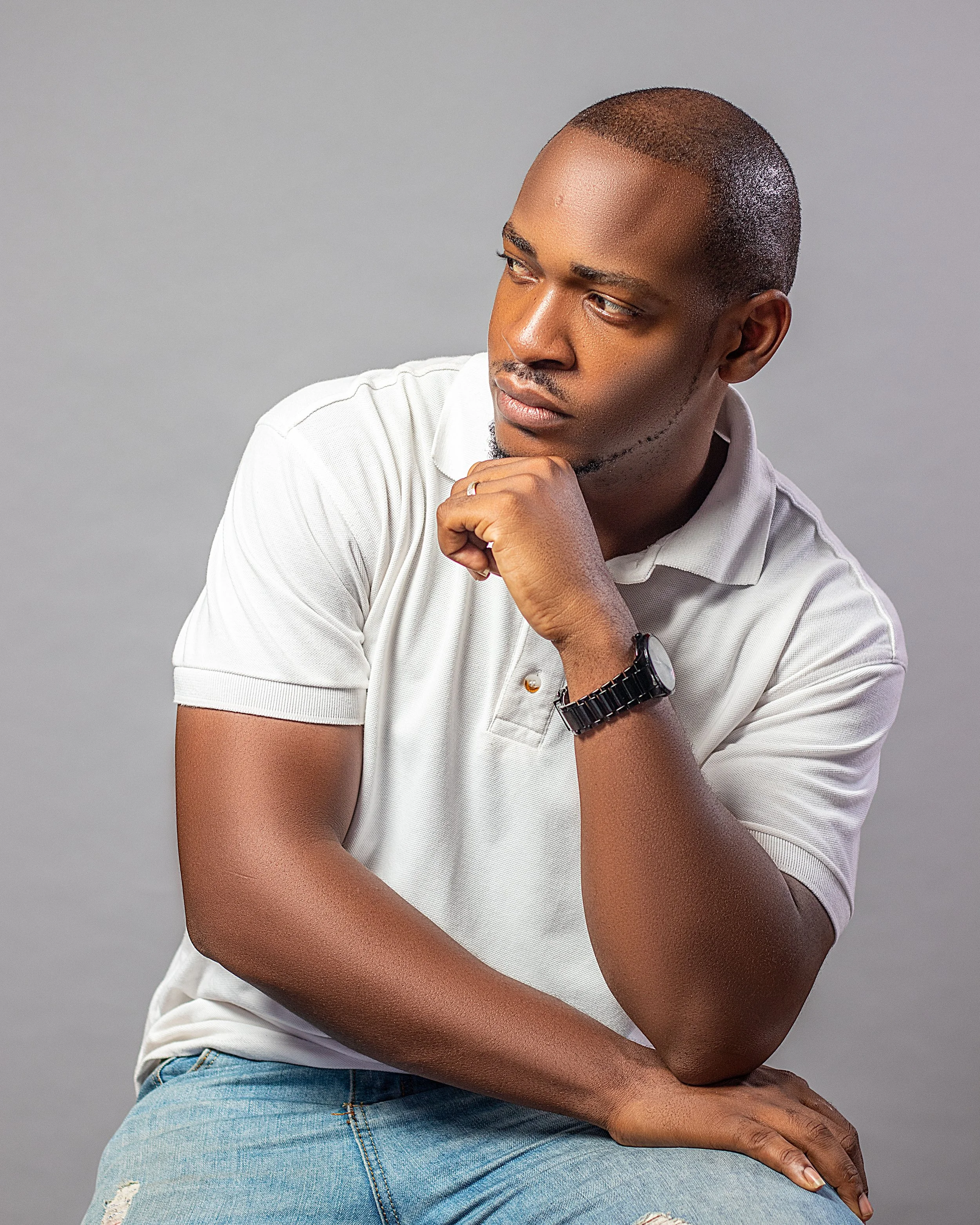 A young man with short hair, wearing a white polo shirt and a black wristwatch, sitting and looking thoughtfully to the side against a plain gray background.