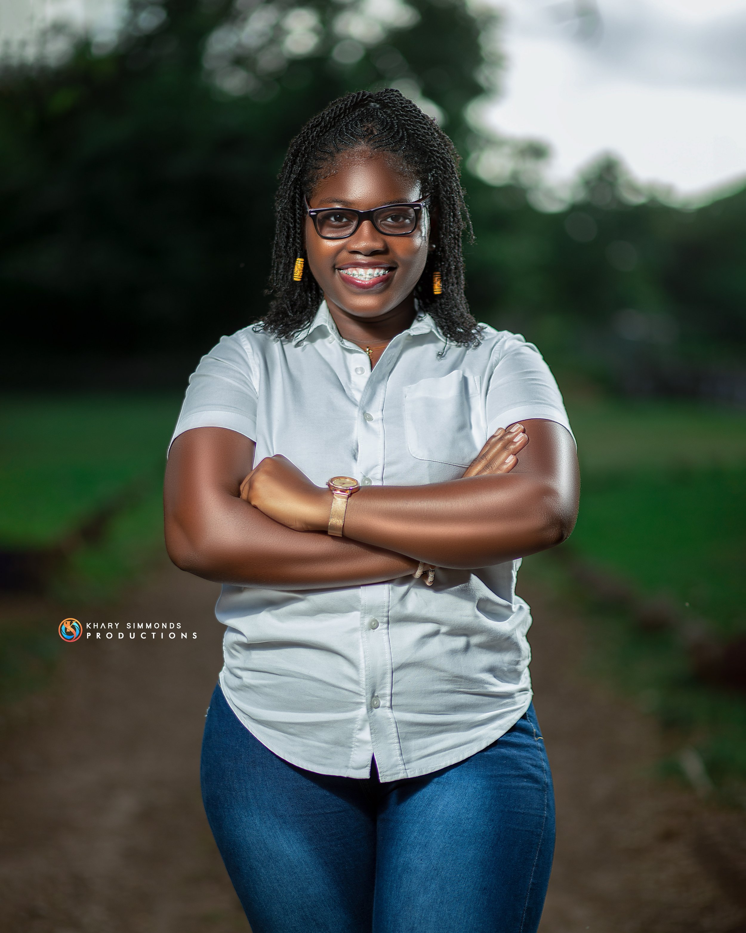 A woman with glasses, braided hair, and a smile, standing outdoors on a path with her arms crossed, wearing a white short-sleeved shirt and blue jeans.