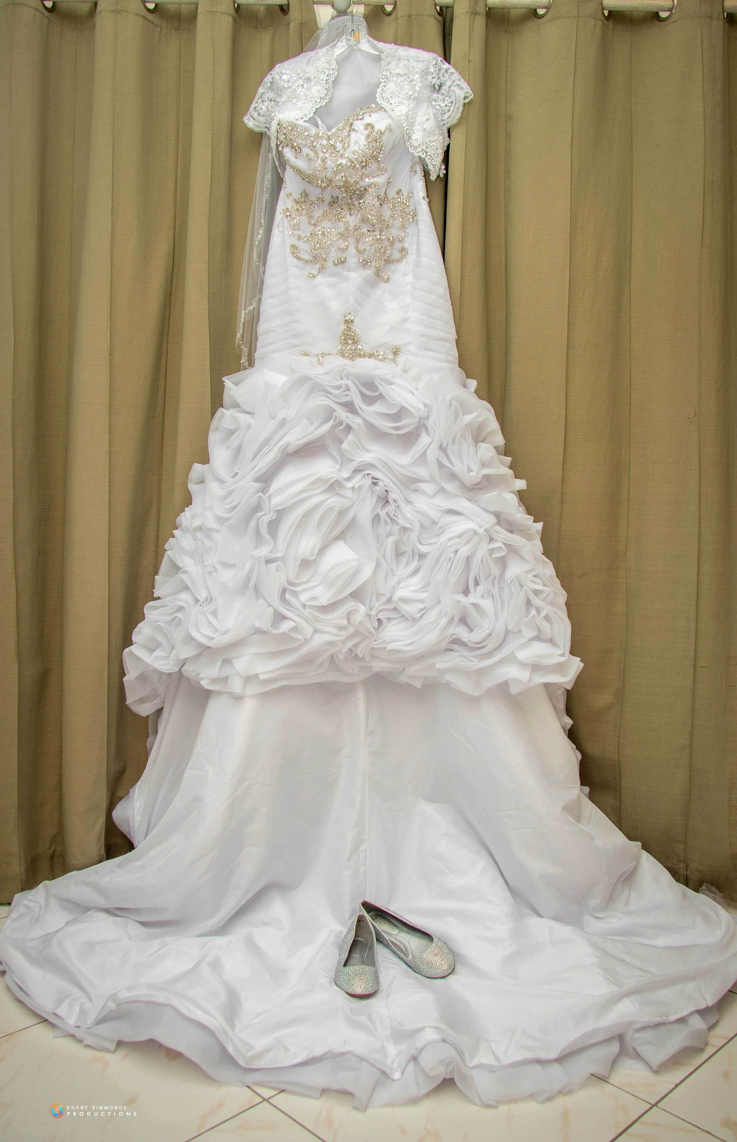 A white wedding dress with intricate embroidery and a ruffled skirt, hanging on a hanger against beige curtains, with a pair of sparkly silver shoes placed at the bottom.