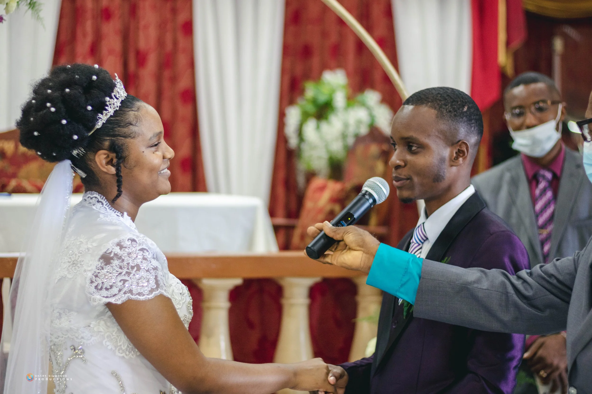 A wedding ceremony with a bride and groom holding hands, facing each other, with a man holding a microphone for vows. The bride is smiling, wearing a white lace dress and a tiara, while the groom is dressed in a purple suit and striped tie. In the ba